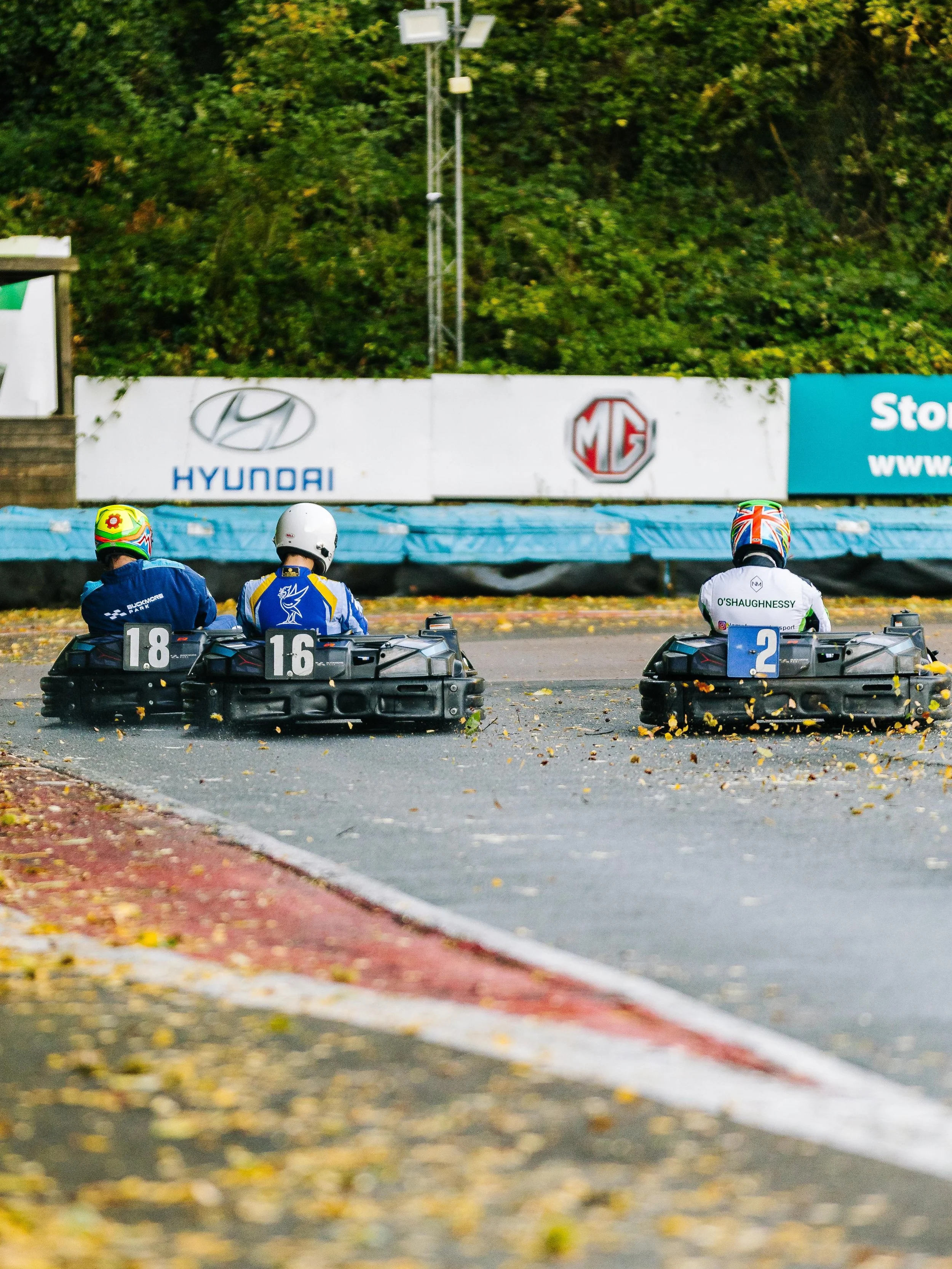 Three individuals racing on go-karts on a race track, with a backdrop of advertisement banners and trees. The drivers are wearing helmets and racing suits, and the go-karts have numbered signs on the back.
