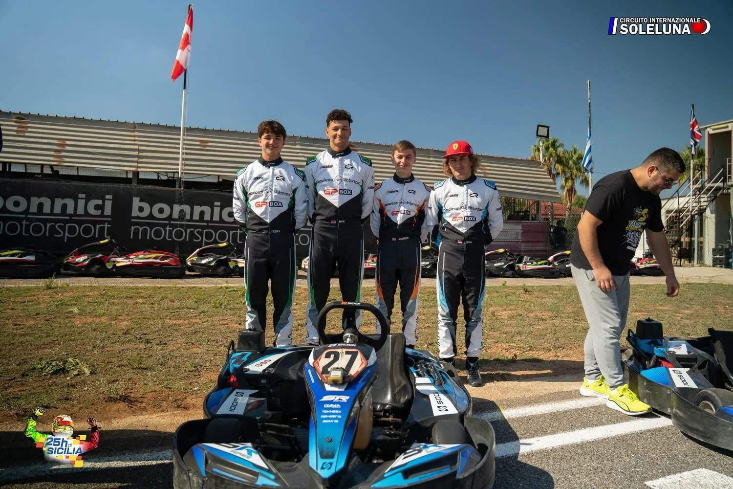 Four race car drivers in racing suits standing on a race track, smiling, with their go-karts behind them. A man on the right is preparing or inspecting a go-kart. The background shows flags, banners, and additional parked go-karts, indicating a racin