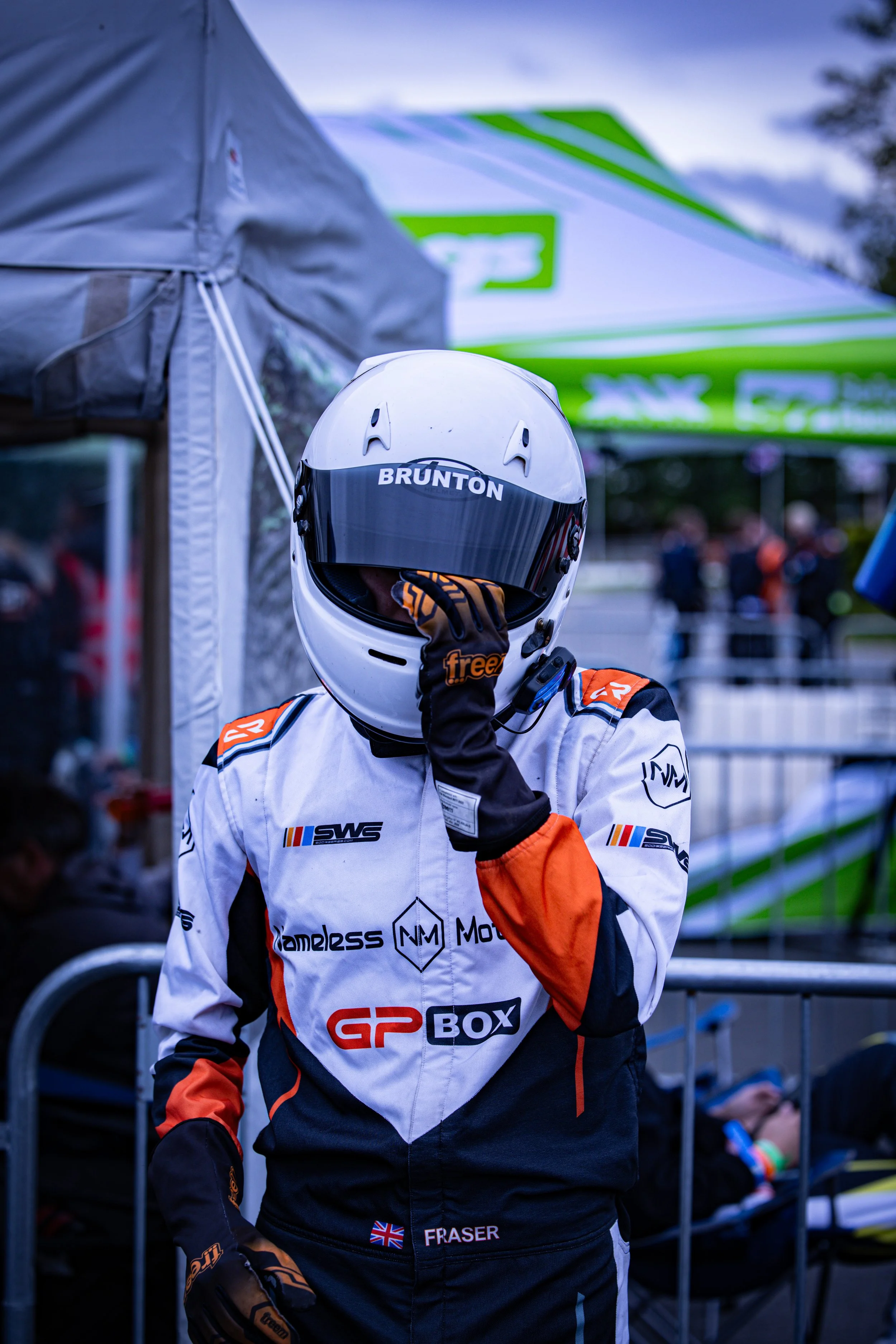Close-up of a person wearing a white racing helmet with a visor marked "BRUNTON," dressed in racing gear. The person appears to be outdoors, possibly preparing for a motorsport event.
