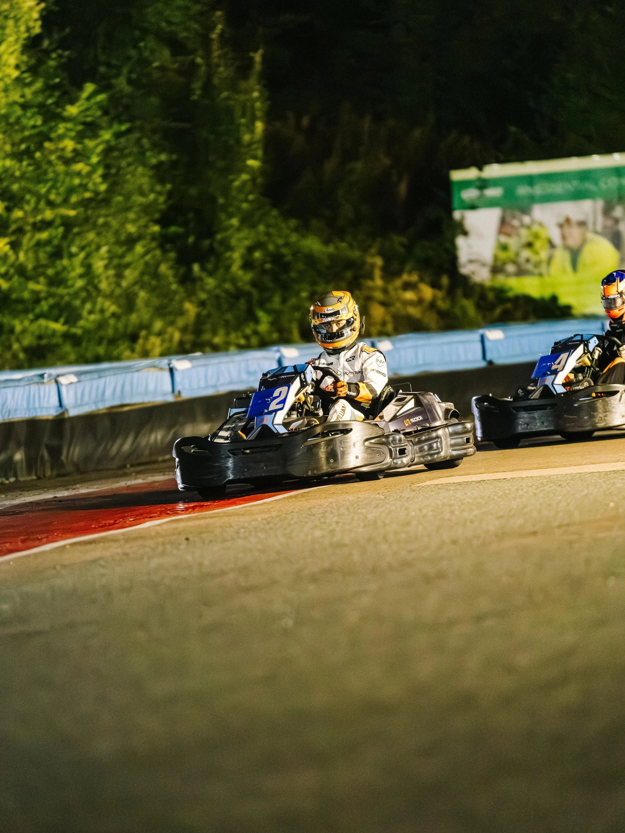 Nighttime go-kart race with two drivers wearing helmets and racing suits, racing on a track with green foliage background.