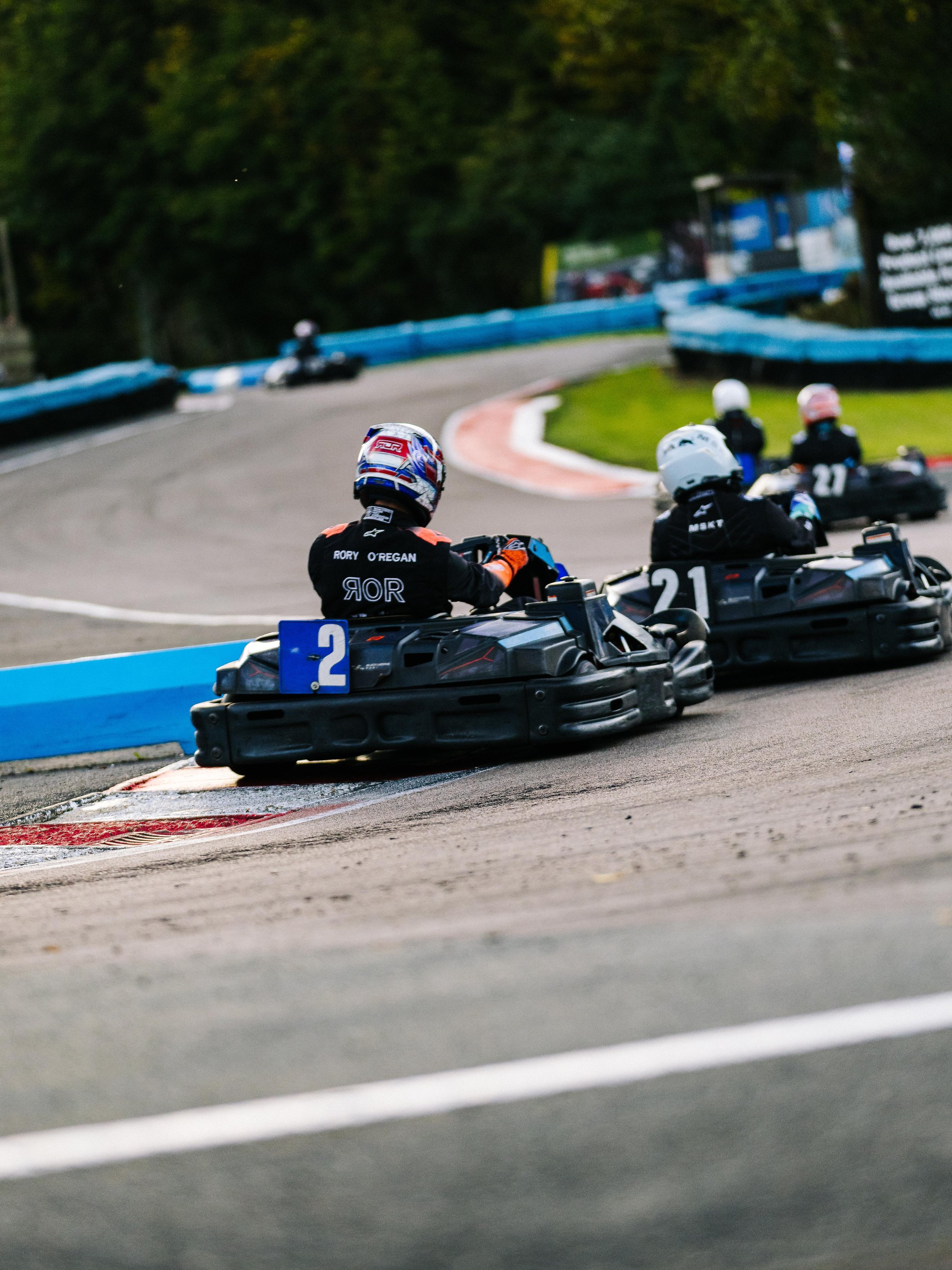 Kart racers on a racing track, with the closest racer in a black suit and white helmet, driving a go-kart numbered 2, followed by another racer in black with a white helmet, and more racers ahead on the track.