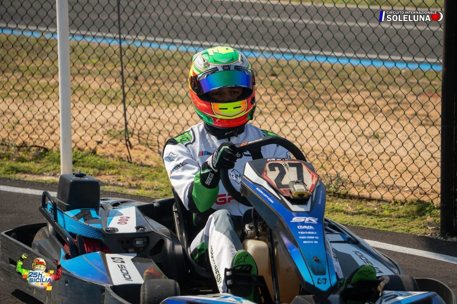 Race car driver in a go-kart wearing a colorful helmet and racing suit, holding the steering wheel on a race track.
