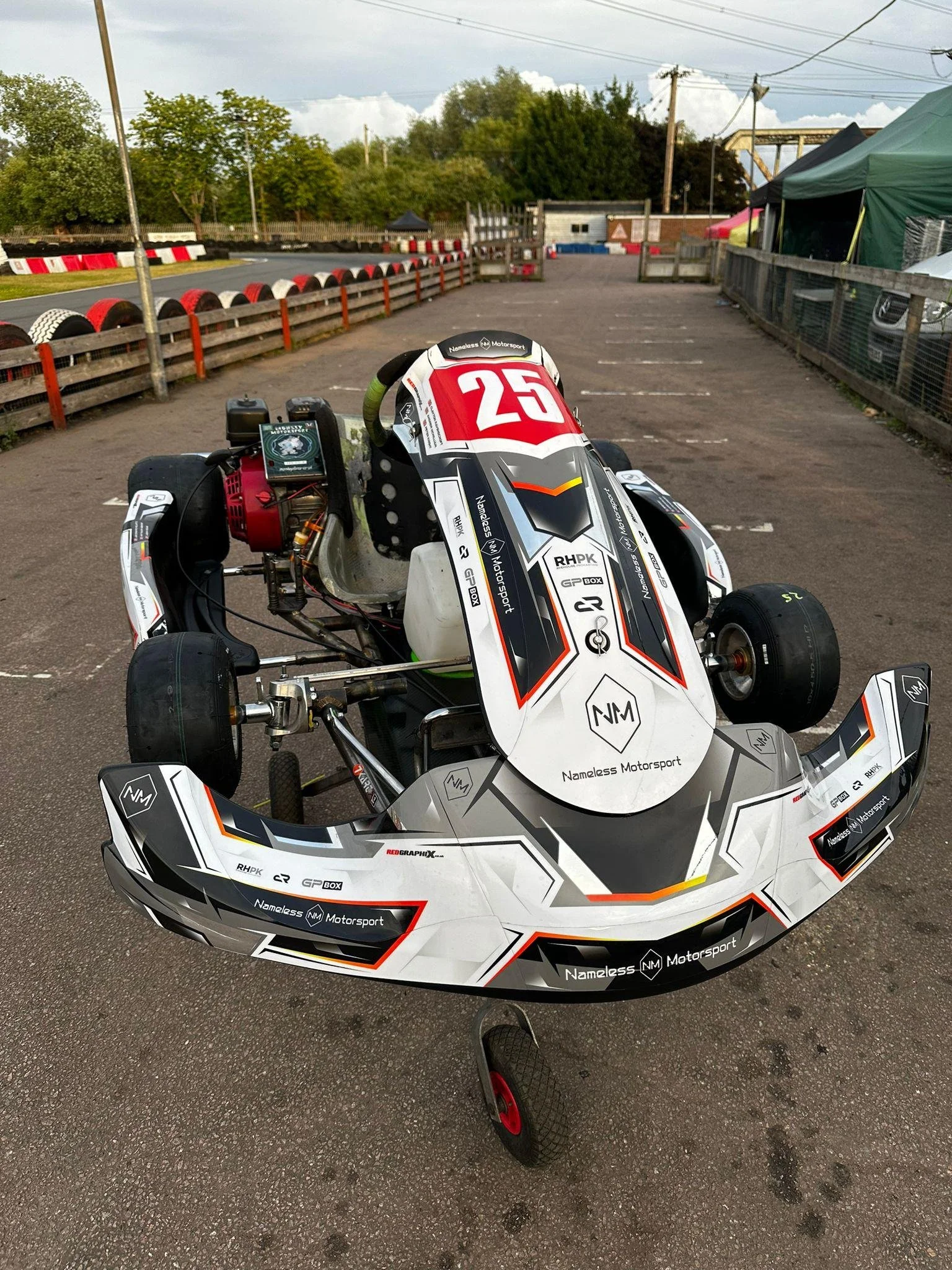 A go-kart with racing decals and the number 25 on the front, parked in an outdoor track area with barriers, tents, and trees in the background.