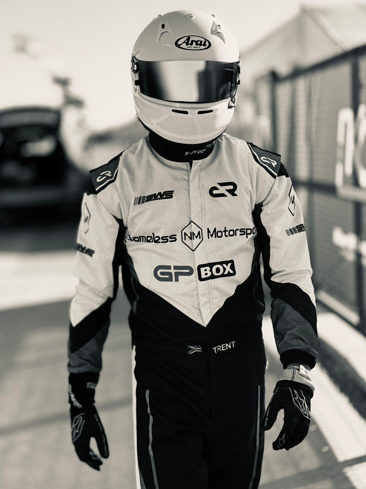 A race car driver in a racing suit and helmet standing in a pit lane, with cars and garages blurred in the background.
