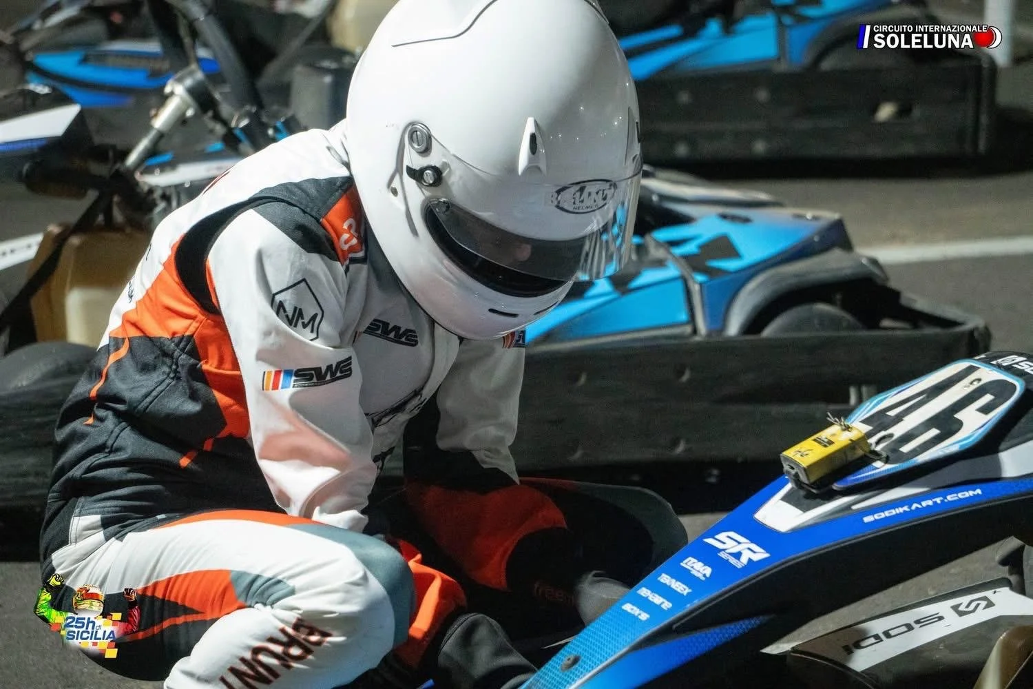 A race car driver wearing a white and orange racing suit and a white helmet getting on a blue race car at night. The background shows other race cars and equipment. There's a logo on the top right corner that says 'Circuito Internazionale Sol Luna'.