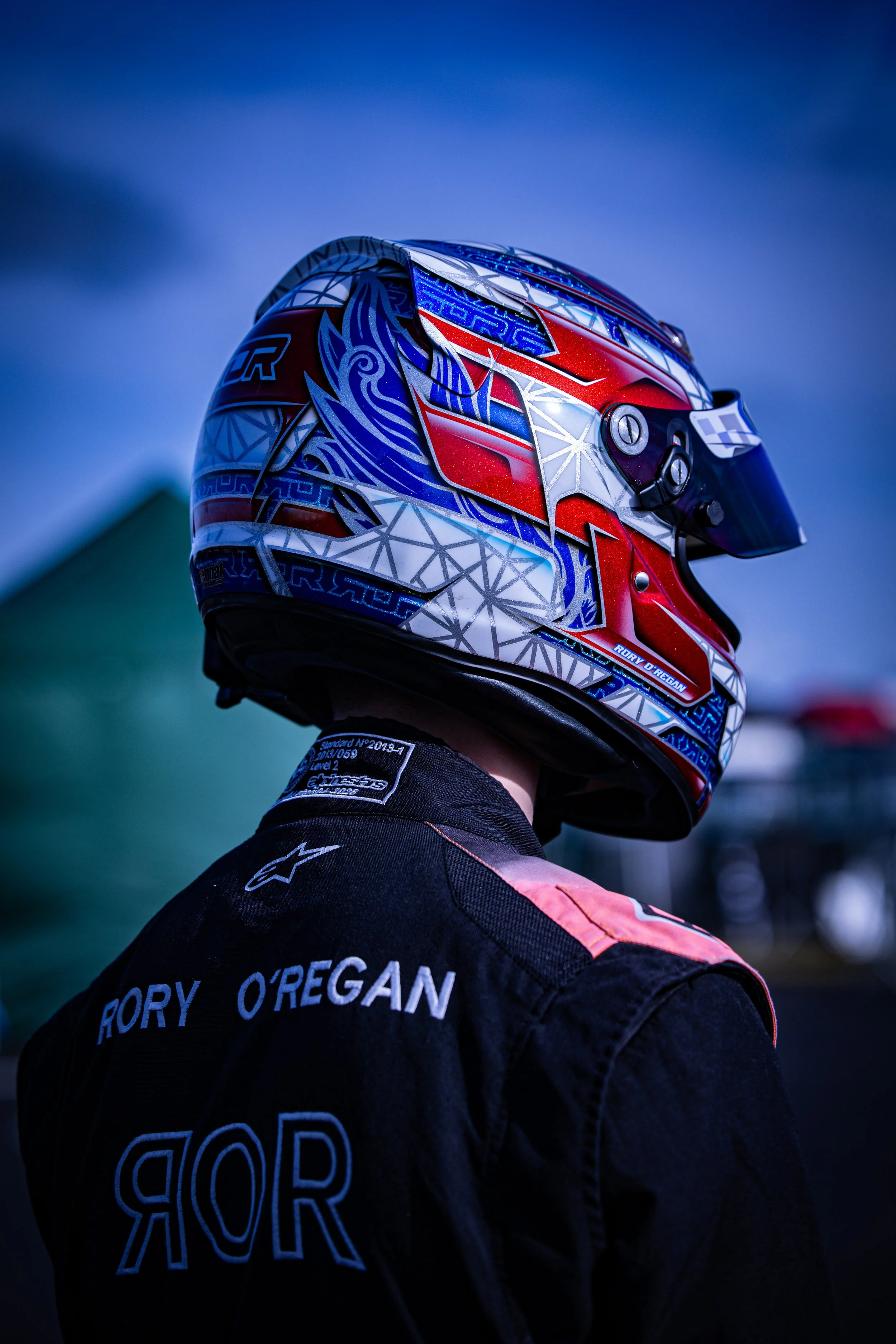 Person wearing a white racing helmet with a dark visor and a black and orange racing suit, standing indoors against a corrugated metal background.