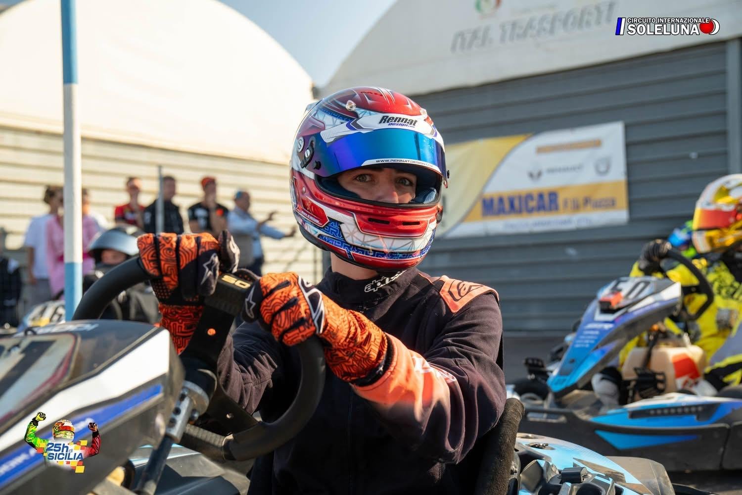 A race car driver in racing gear and a blue, red, white helmet sitting in a go-kart, with other racers and spectators in the background at a racing event.