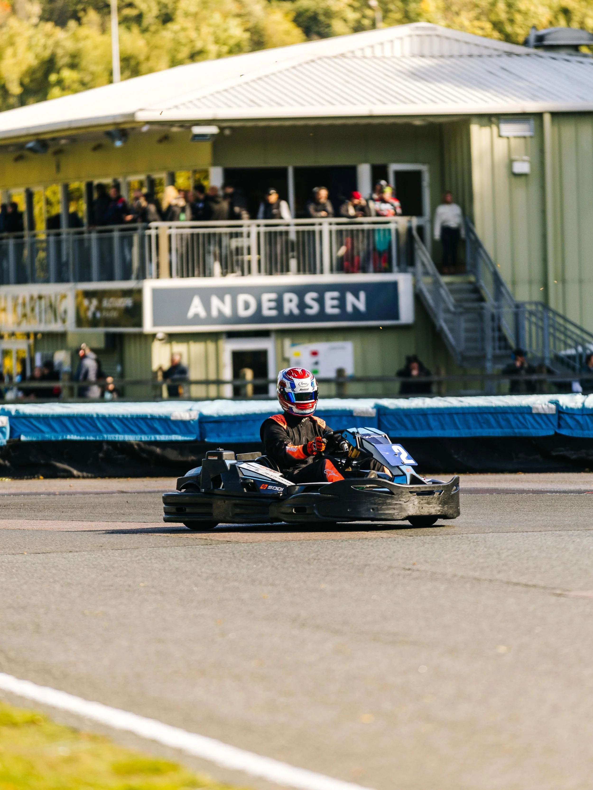 A person driving a go-kart on a race track with a green building and spectators in the background.