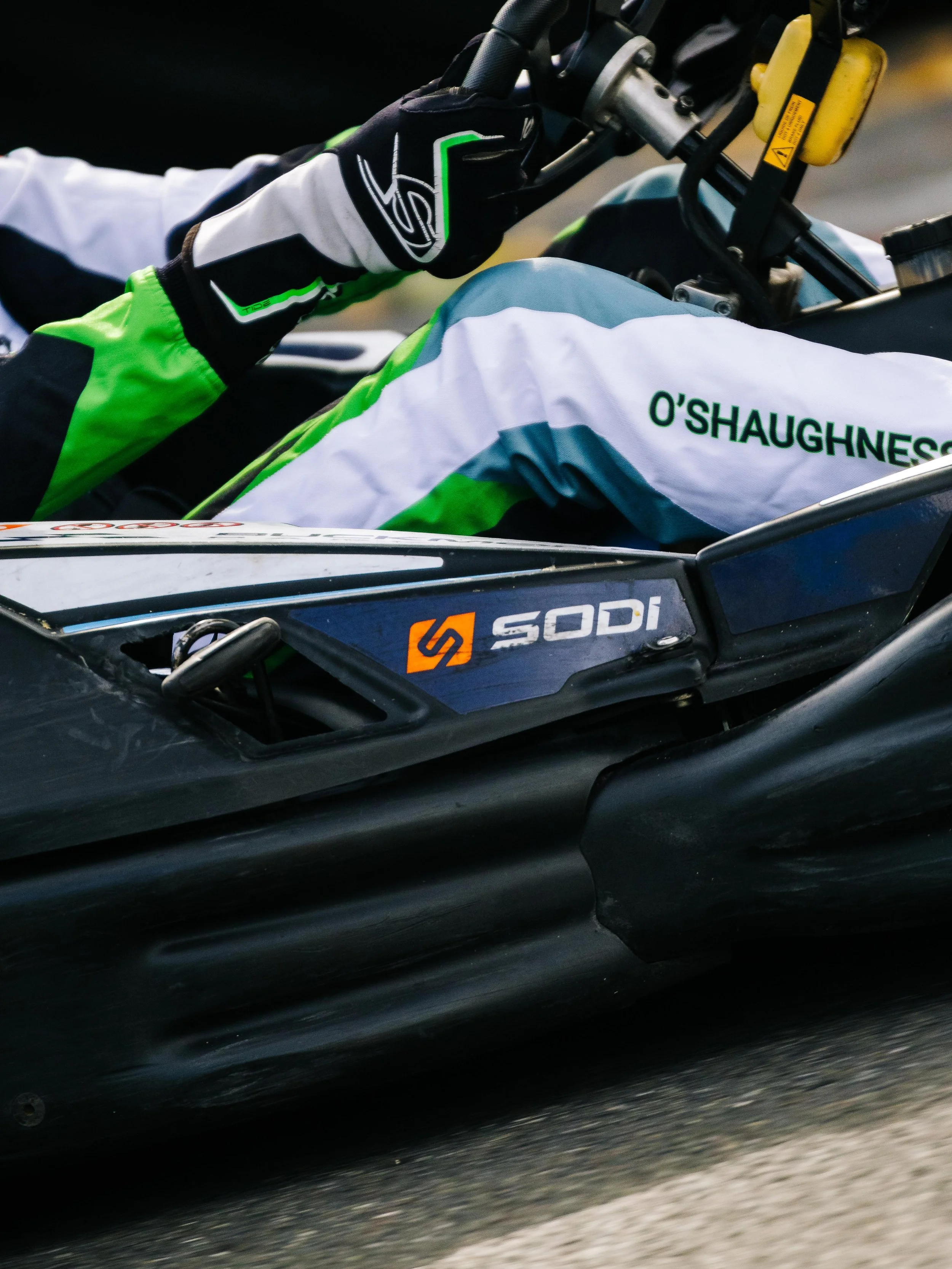 Close-up of a race car driver gripping the steering wheel, wearing a racing suit with 'O'Shaughnessy' on it, in a black and blue race car with Sodi branding.