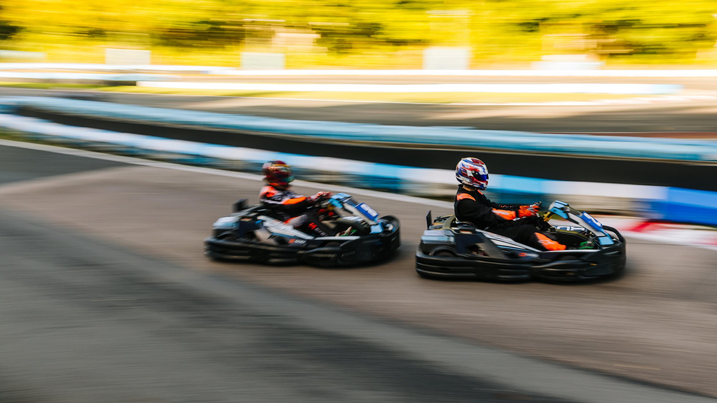 Two people in go-karts racing on a track, blurred background indicating high speed with autumn-colored trees in the background.