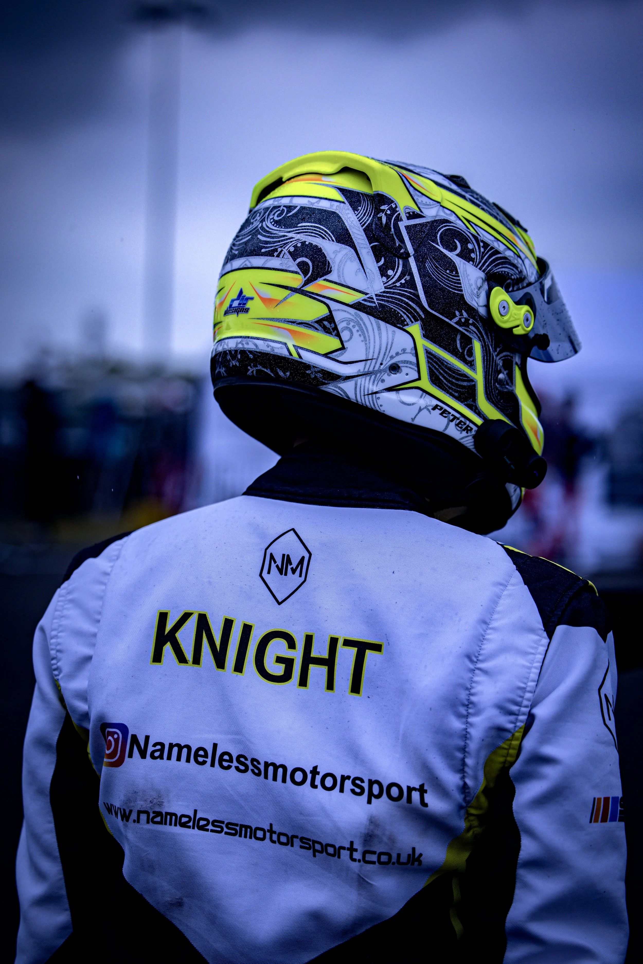 Race car driver wearing a white helmet and racing suit with a prominent logo, standing in a pit area.