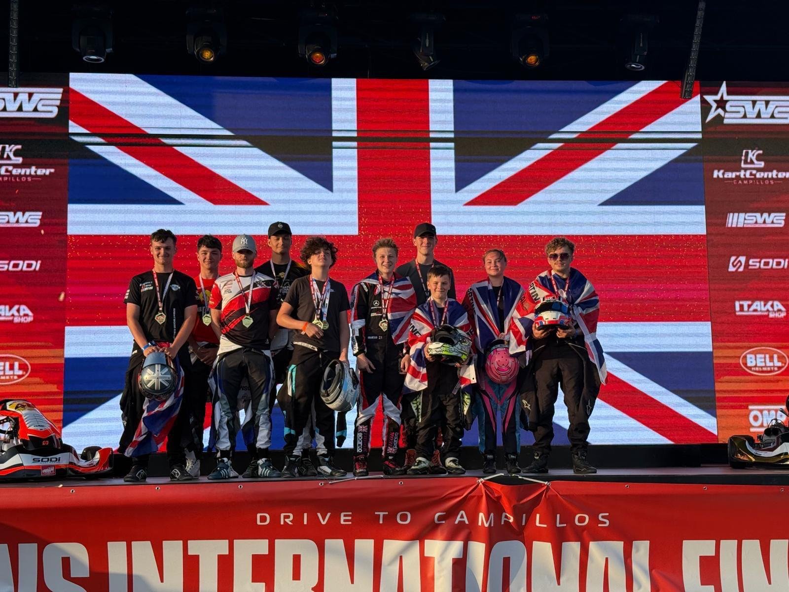 A group of race car drivers standing on a podium, holding helmets and medals, with a British flag backdrop and a red banner that says 'Drive to Campillos' and 'International.'