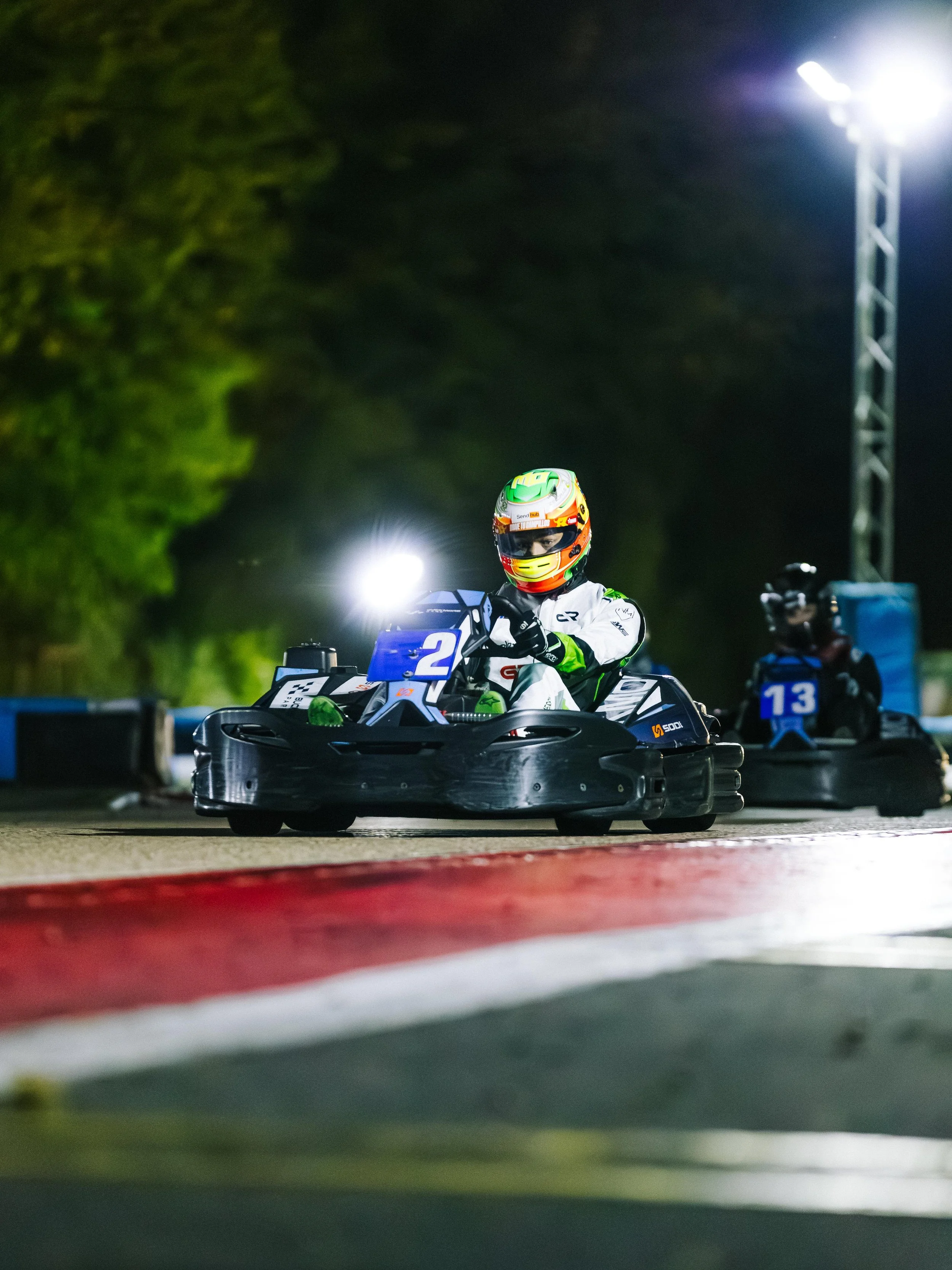 Nighttime go-kart race featuring a driver in a white and green suit and helmet, driving a black go-kart with the number 2, illuminated by bright lights on a race track with red, white, and black curbing.