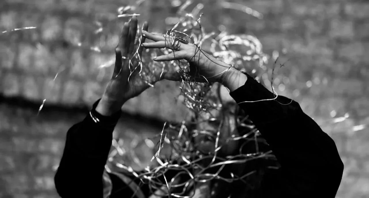 A person with black clothing and bracelets on their wrist is tangled in strands of thread or wire, with their hands raised and intertwined with the strands, against a blurred brick wall background.
