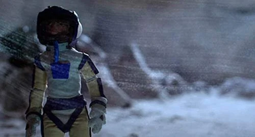 Astronaut in space suit standing outdoors with a view of the ocean or a large body of water.