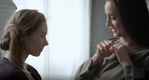 Two women face each other indoors, one smiles while helping the other with a jacket or coat.