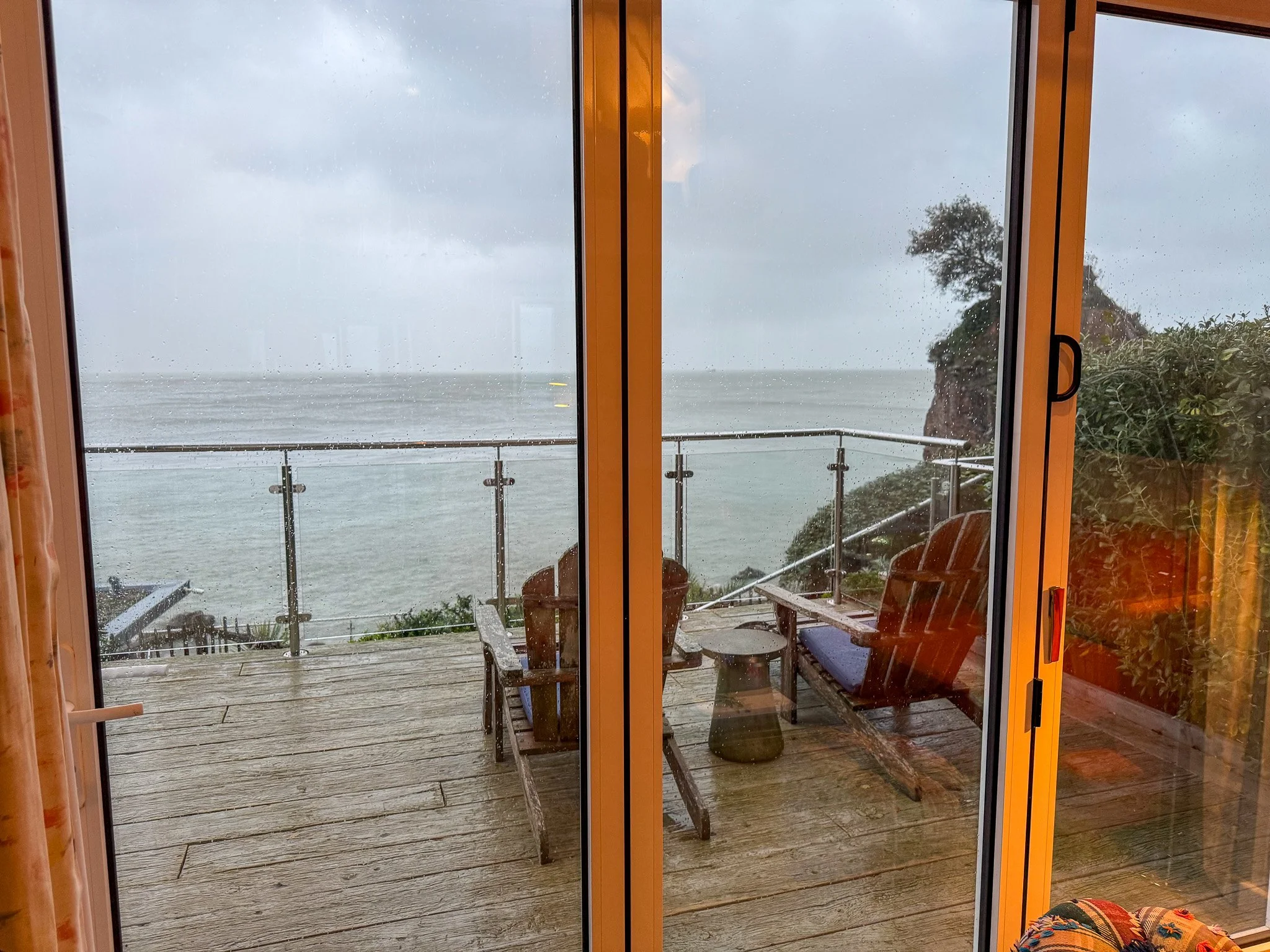 View from the lounge of a Cary Arms beach hut looking out to a private decking area with loungers and sea view in Torquay, Devon