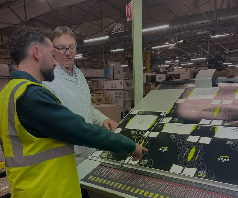 Deux hommes, un portant un gilet de sécurité, examinent une machine d'emballage ou d'impression dans une usine.