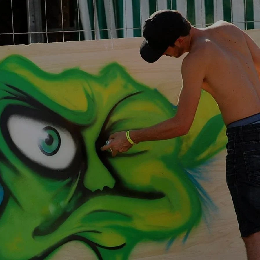 Jeune homme en train de faire du graffiti sur un panneau en bois, représentant un visage colore en vert avec un œil grand et un air fâché, portant une casquette noire et un bracelet jaune.