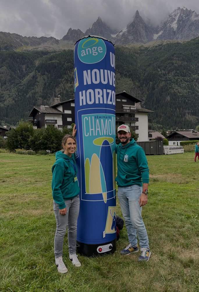Deux personnes posent à côté d'une grande colonne gonflable de couleur bleue avec des inscriptions en français, dans un paysage de montagne avec des maisons et des arbres.