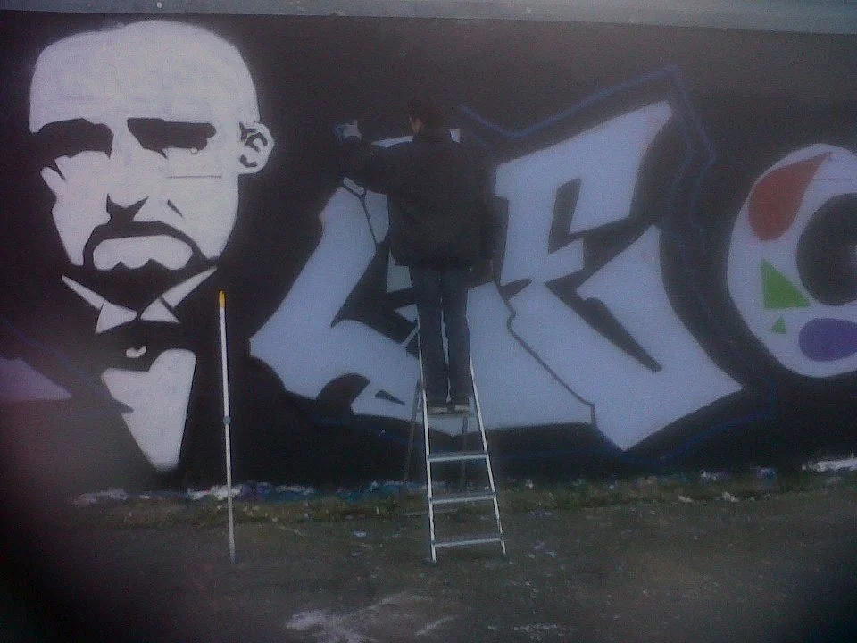 Un artiste peint une fresque murale représentant une figure en noir et blanc, ressemblant à une image de style soviétique, à côté d'un graffiti coloré, vue de dos sur une échelle.
