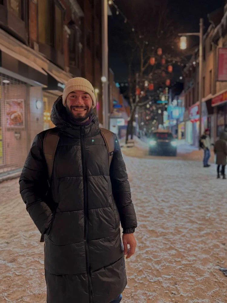 Un homme souriant portant un manteau noir, un bonnet beige et un sac à dos, debout dans une rue urbaine la nuit, avec des lanternes suspendues et des bâtiments éclairés, au sol de la neige.