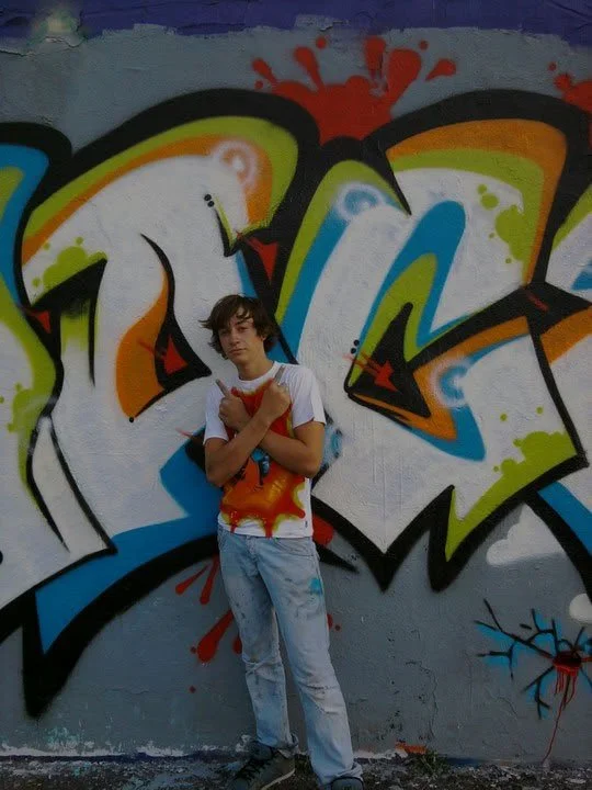 Jeune garçon posant devant un mur avec graffiti coloré, portant un T-shirt blanc avec un motif orange.