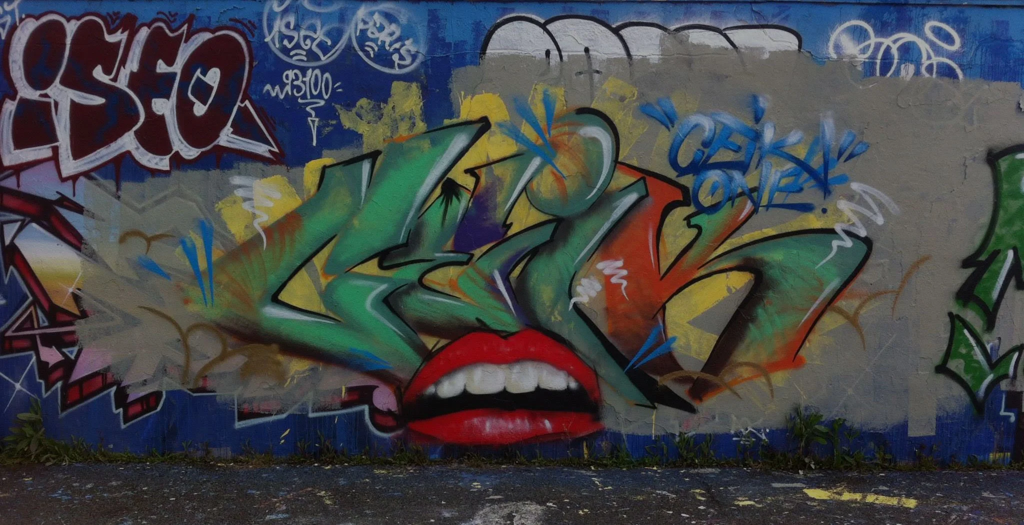 Graffiti coloré représentant un visage avec des lèvres rouges et un maquillage artistique. Titre en graffiti dans des tons de vert, orange, bleu et jaune, avec d'autres motifs et tags autour.