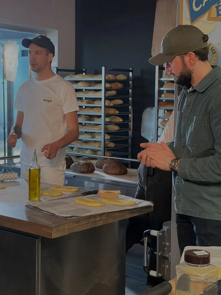 Deux hommes en train de préparer et de parler dans une boulangerie. Il y a des pains frais sur des étagères derrière eux et des pâtons de pâte à pain sur le plan de travail.