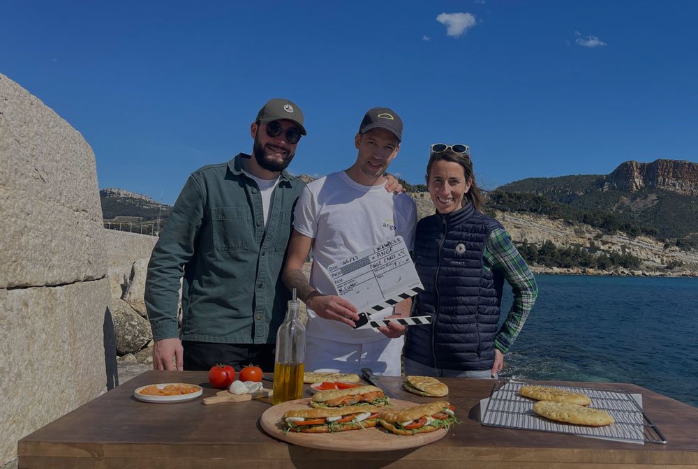 Trois personnes posent devant une table avec des sandwiches et des tomates, à l'extérieur près de la mer, avec un ciel bleu.