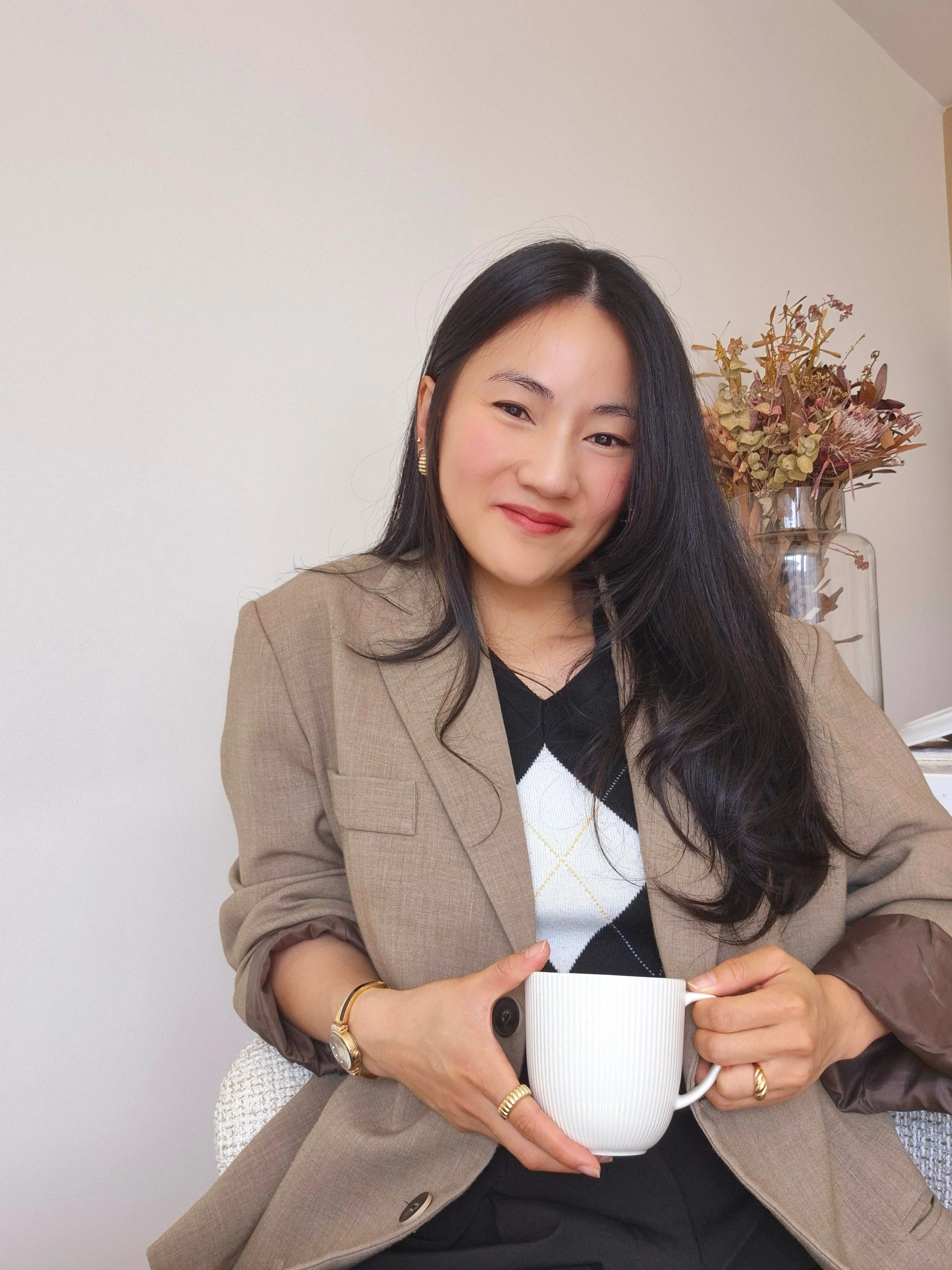 Woman in a beige blazer holding a white mug with a neutral background and dried flowers.