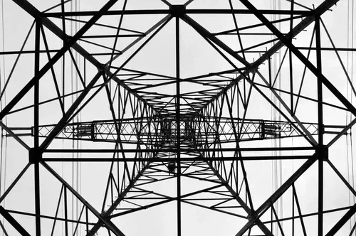 Looking up at the understructure of a tall metal electricity transmission tower with a lattice framework and power lines running through it, in black and white.