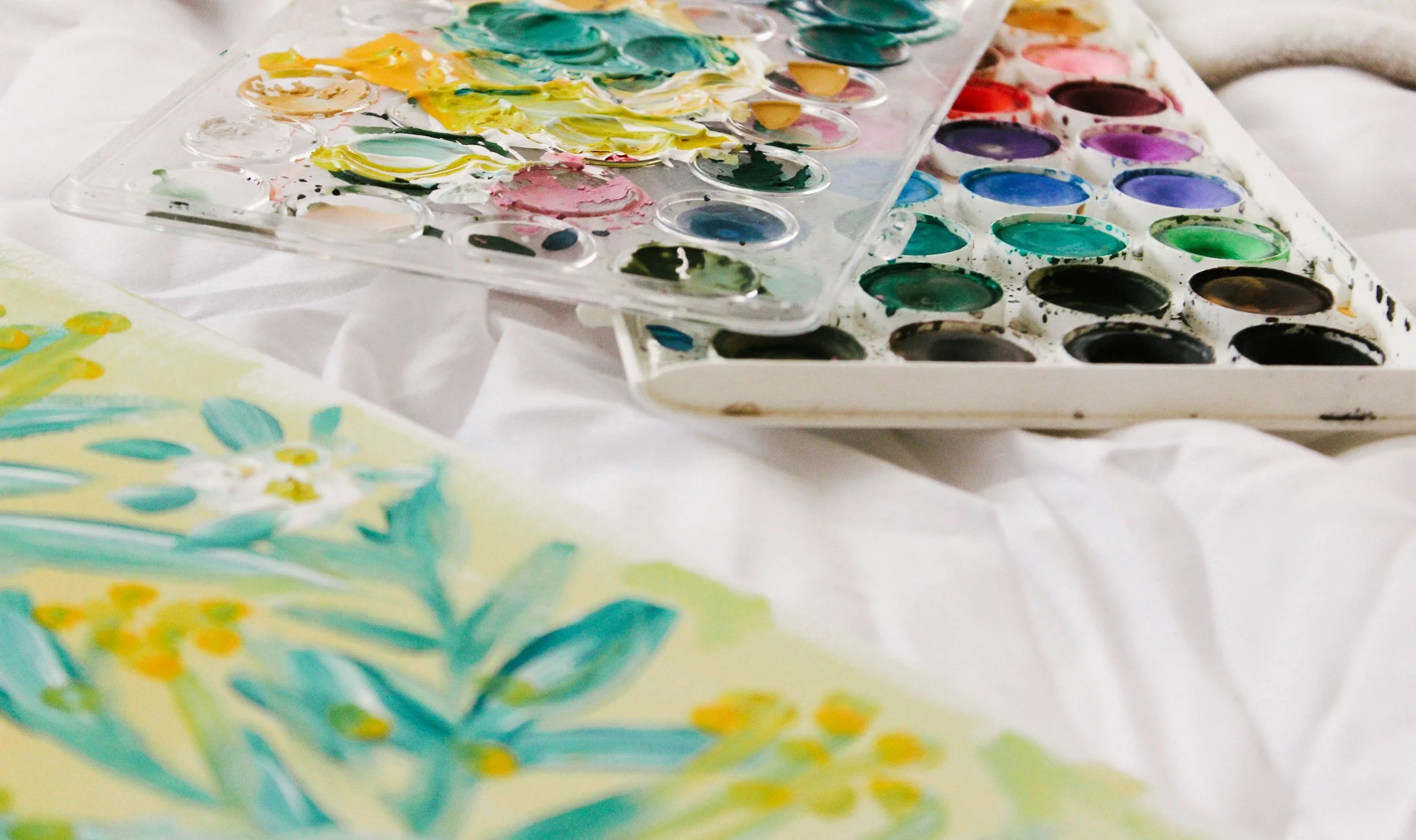 Watercolor paint palette with various colors and a partially painted floral artwork with yellow, teal, and white flowers on paper.