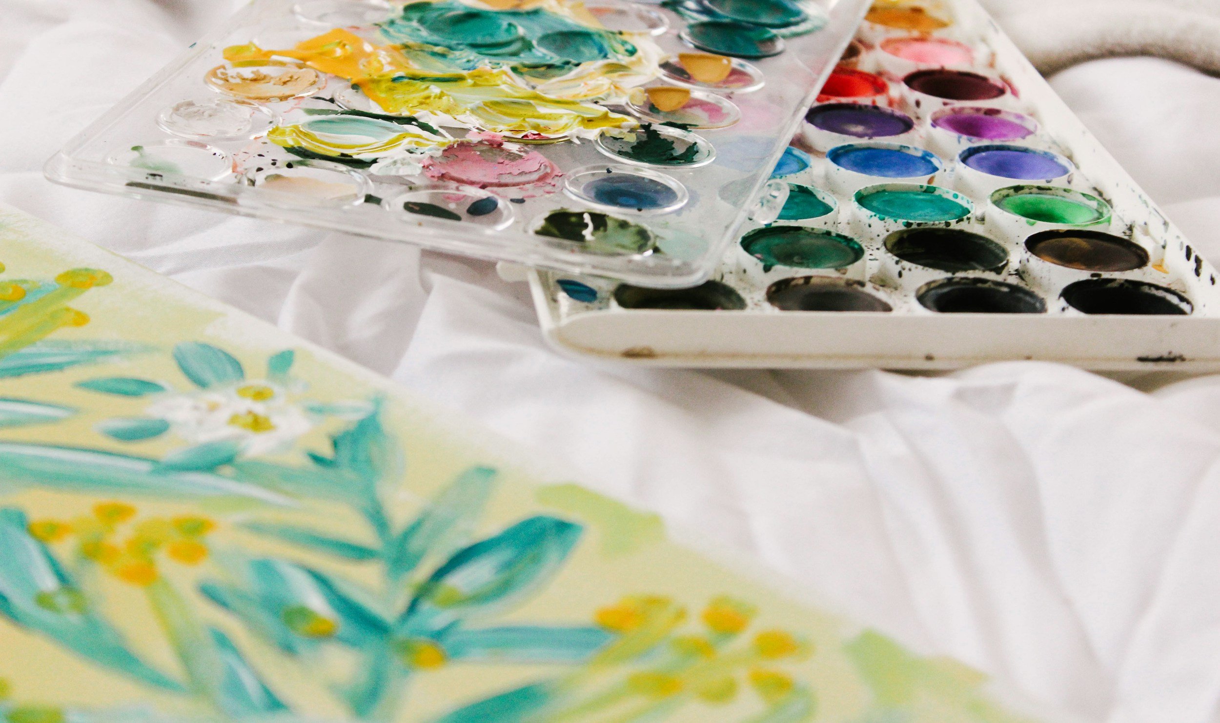 Paint palettes with various colors and a partially painted artwork featuring floral designs on paper.