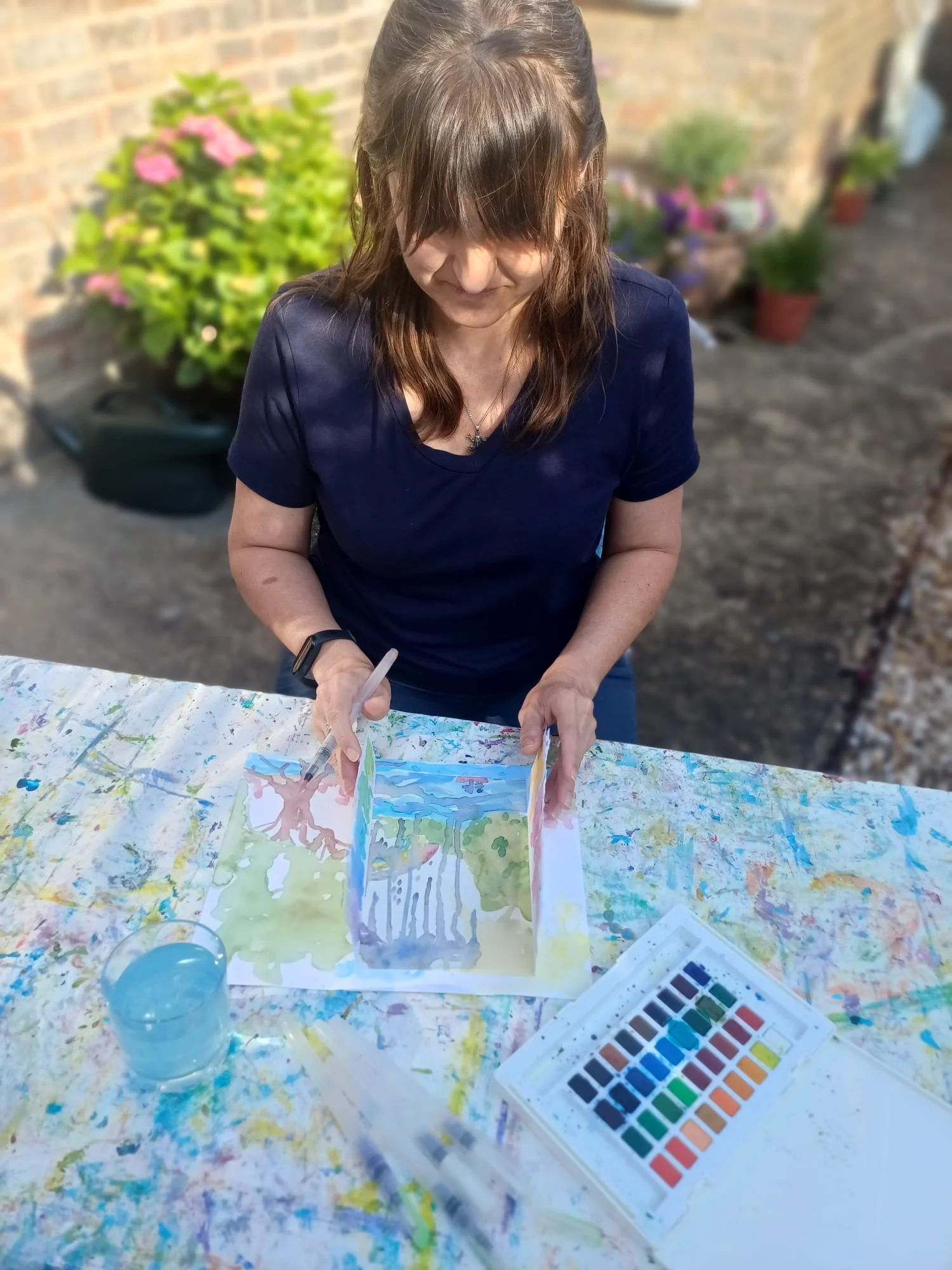 Art Therapist Janaline. Woman with brown hair in a ponytail, wearing a black shirt, painting watercolor art outdoors on a table with water and paint set, with plants and a brick wall in the background.