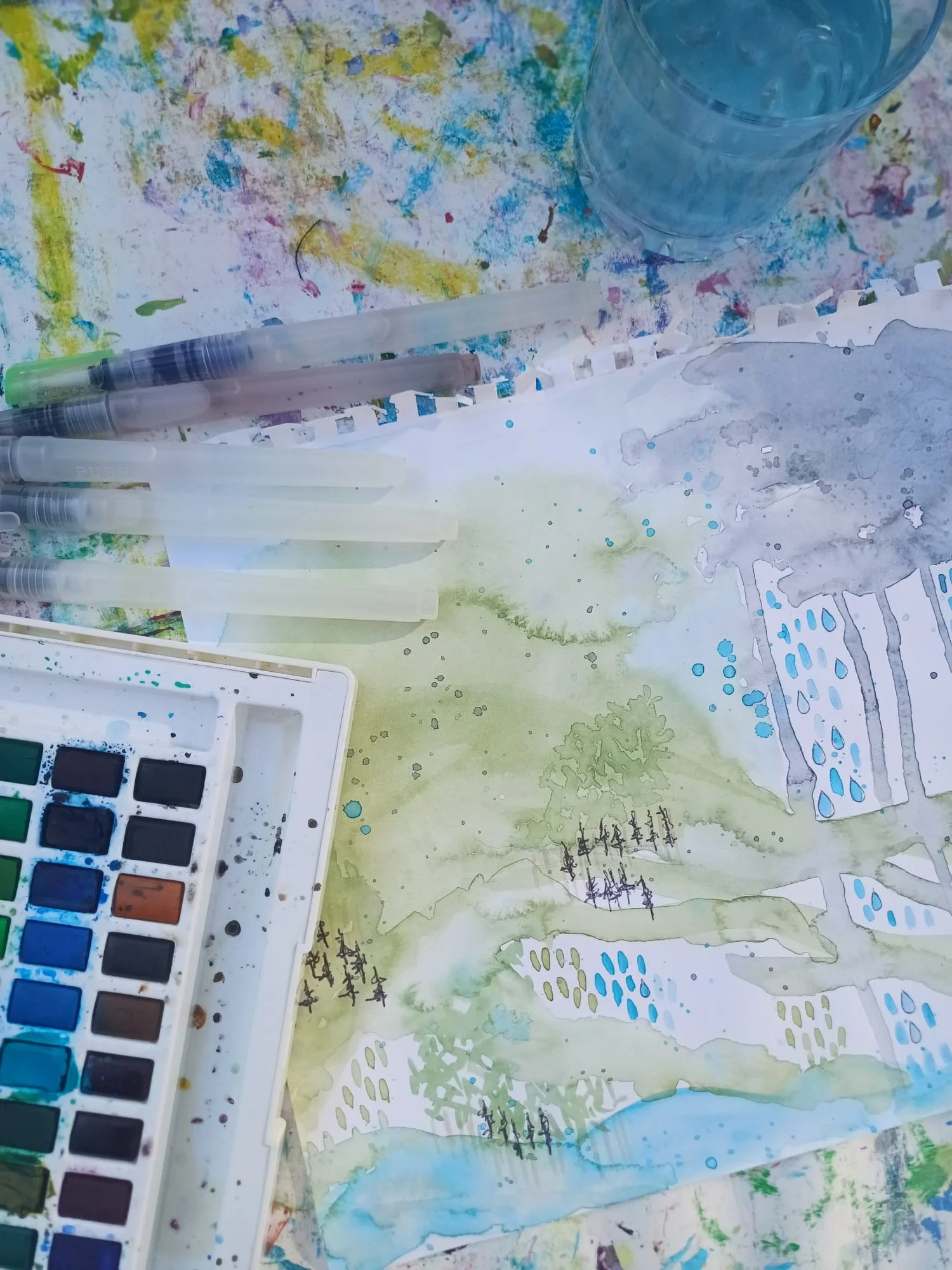 Art supplies including watercolor paints, brushes, water cup, pens, and watercolor paper with mixed paint and ink markings.