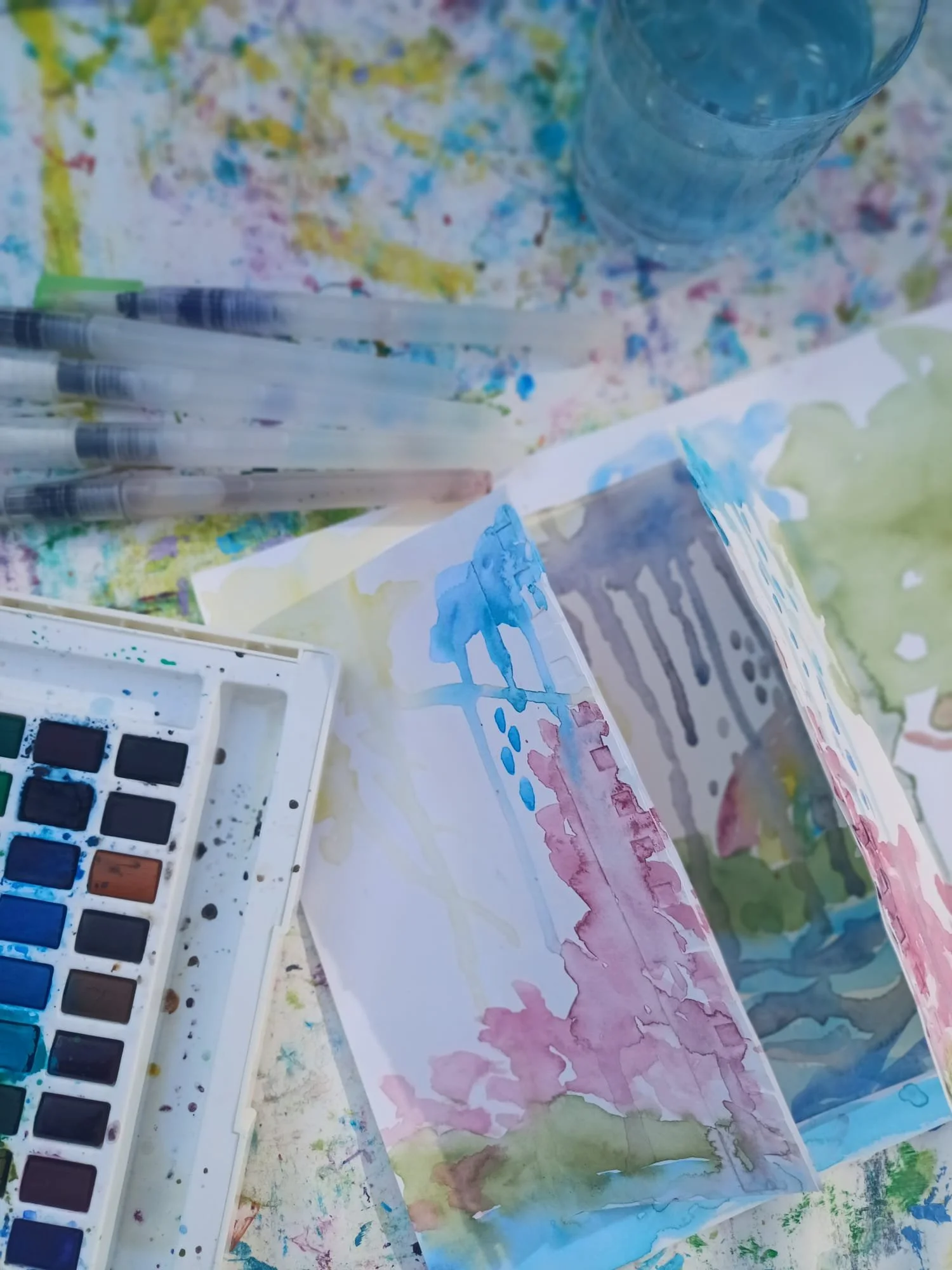 Paint supplies on a splattered table, including watercolor paints, pens, and paper with watercolor paintings featuring blue, pink, and green hues.