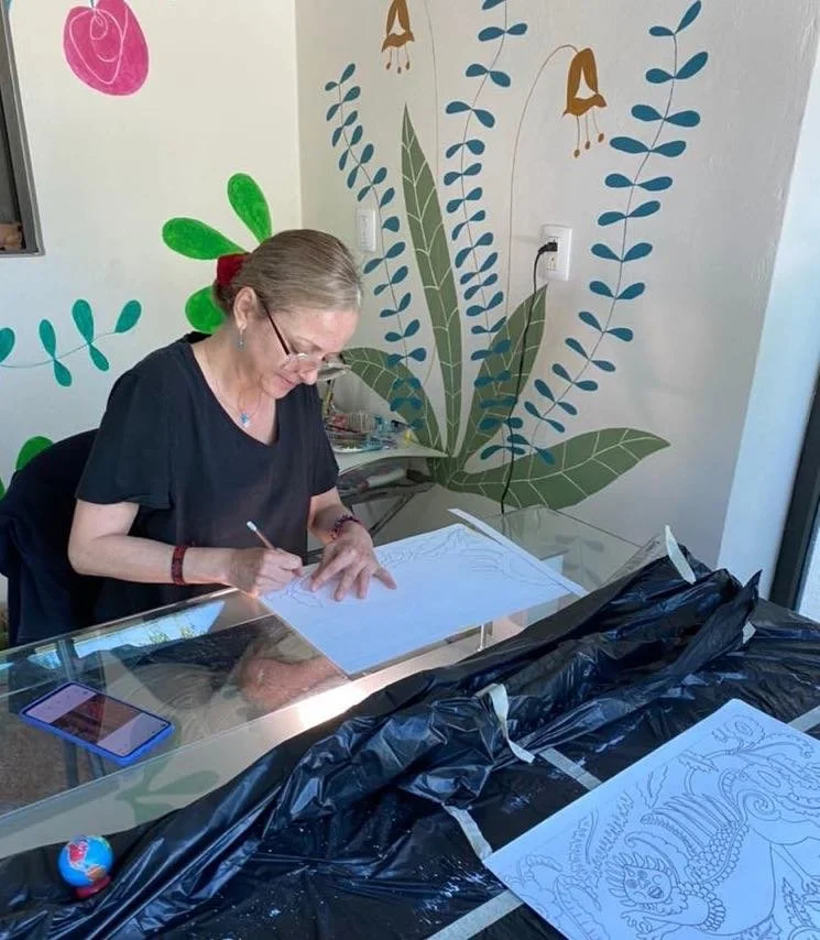 A woman with glasses and a red hair tie is sitting at a glass table, sketching on white paper with a pencil. She is surrounded by art supplies and has a smartphone nearby. The wall behind her features a colorful mural with large green leaves, blue leaves, and stylized brown and gold flowers.