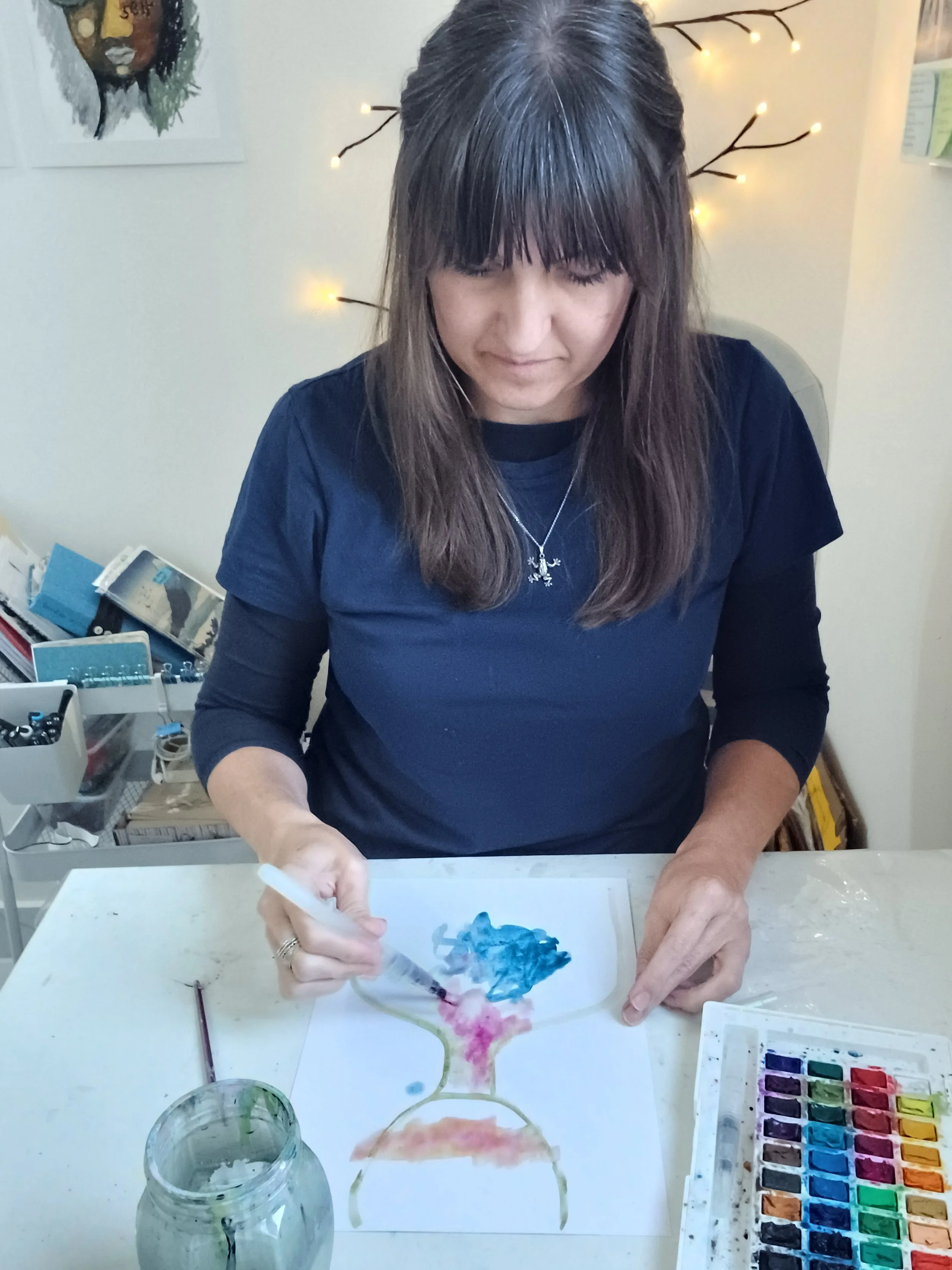 Janaline, therapeutic art facilitator , is sitting at a table creating watercolor art. She is holding a figurative brush with blue and pink watercolors on her paper.  There are various watercolor paints and art supplies on the table.