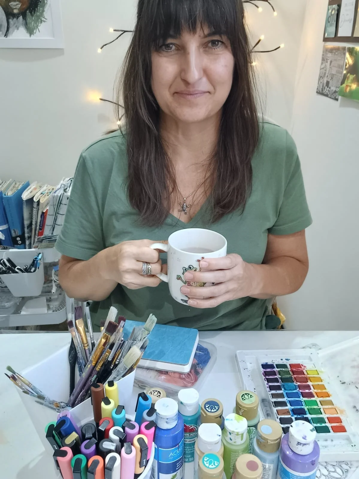 Janaline, therapeutic art facilitator supporting expat women through creative emotional support holding a coffee mug, surrounded by art supplies on a table, including paint brushes, watercolor paints, and markers.