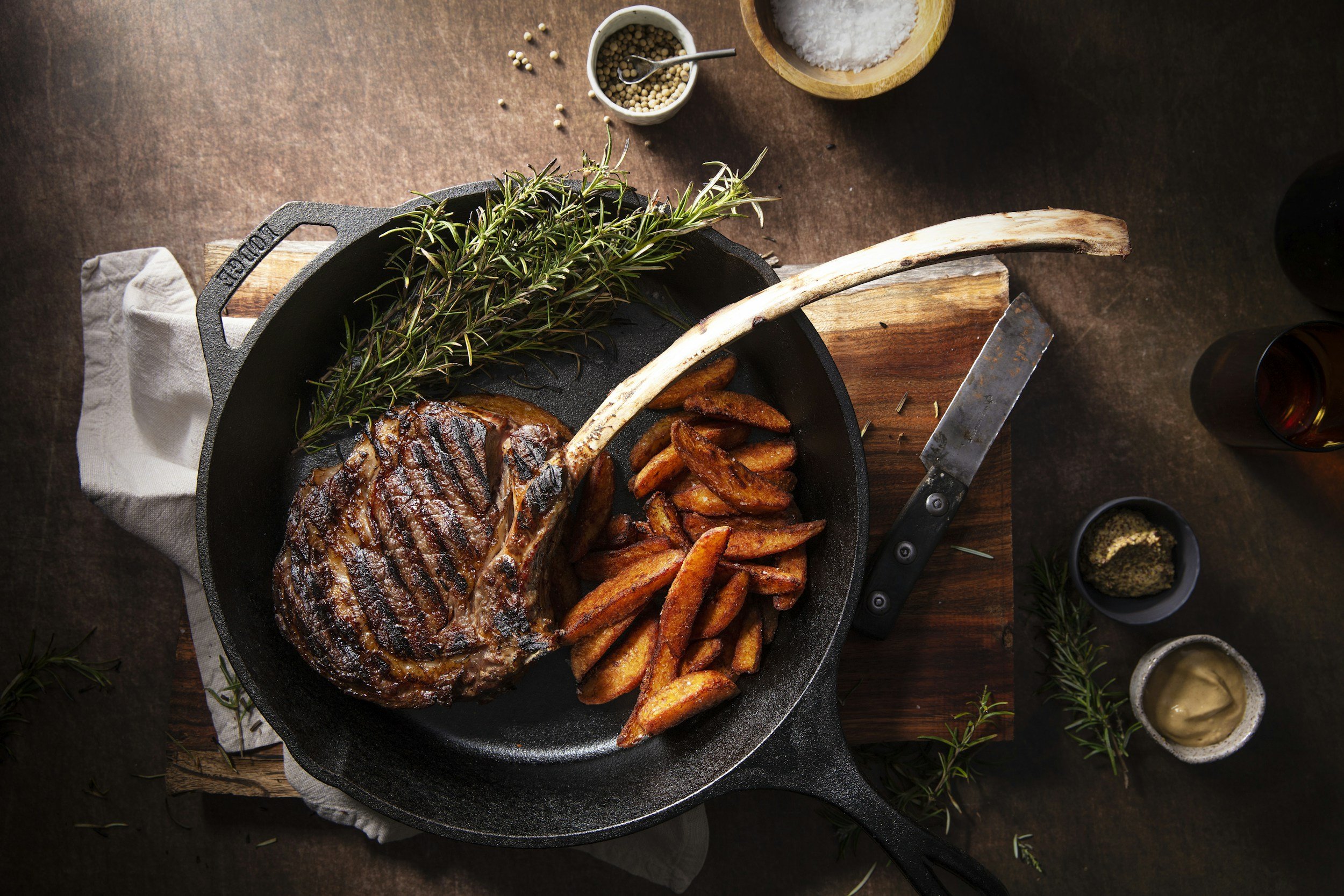 A grilled steak with grill marks served in a cast iron skillet, accompanied by crispy potato wedges and a sprig of fresh rosemary. The skillet rests on a wooden cutting board, with small bowls of salt, peppercorns, mustard, and mustard seeds nearby, on a dark wooden table.