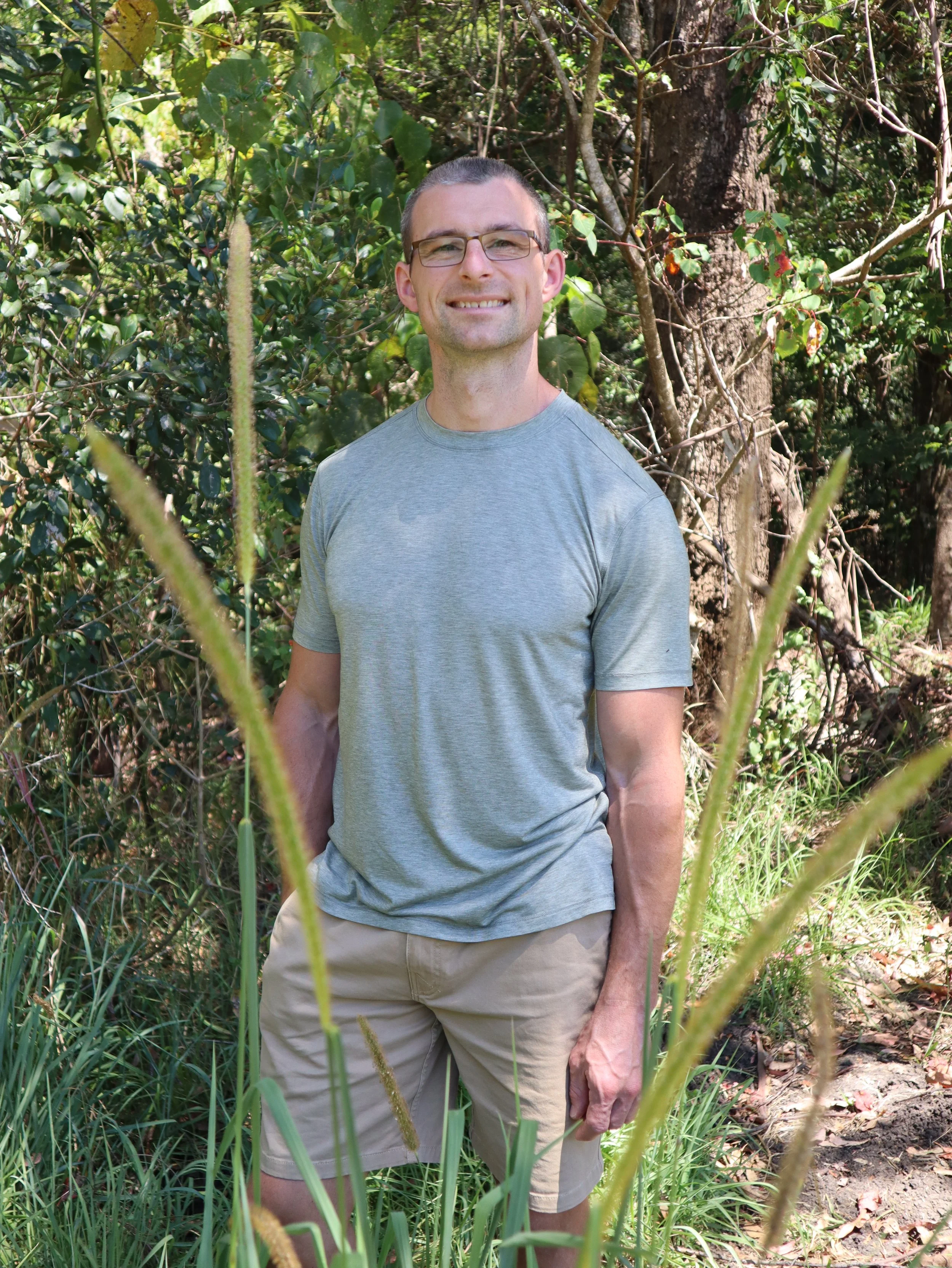A man with short hair and glasses smiling in a wooded outdoor area, wearing a gray t-shirt and khaki shorts, surrounded by tall grass and trees.