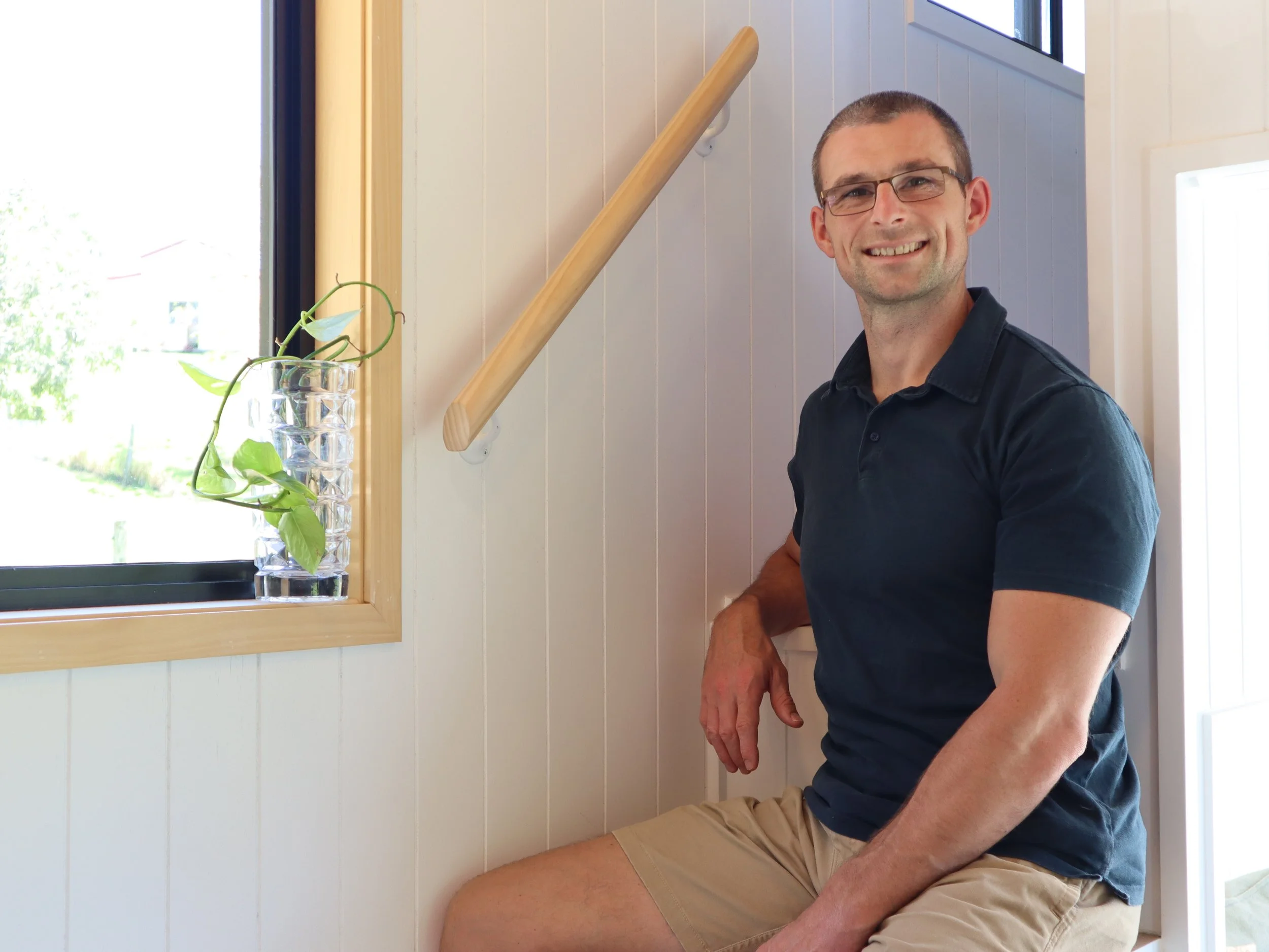 A man sitting by a window, smiling, wearing glasses, a dark blue polo shirt, and tan shorts, with a wooden handrail and a small glass vase with a green leafy plant next to him.