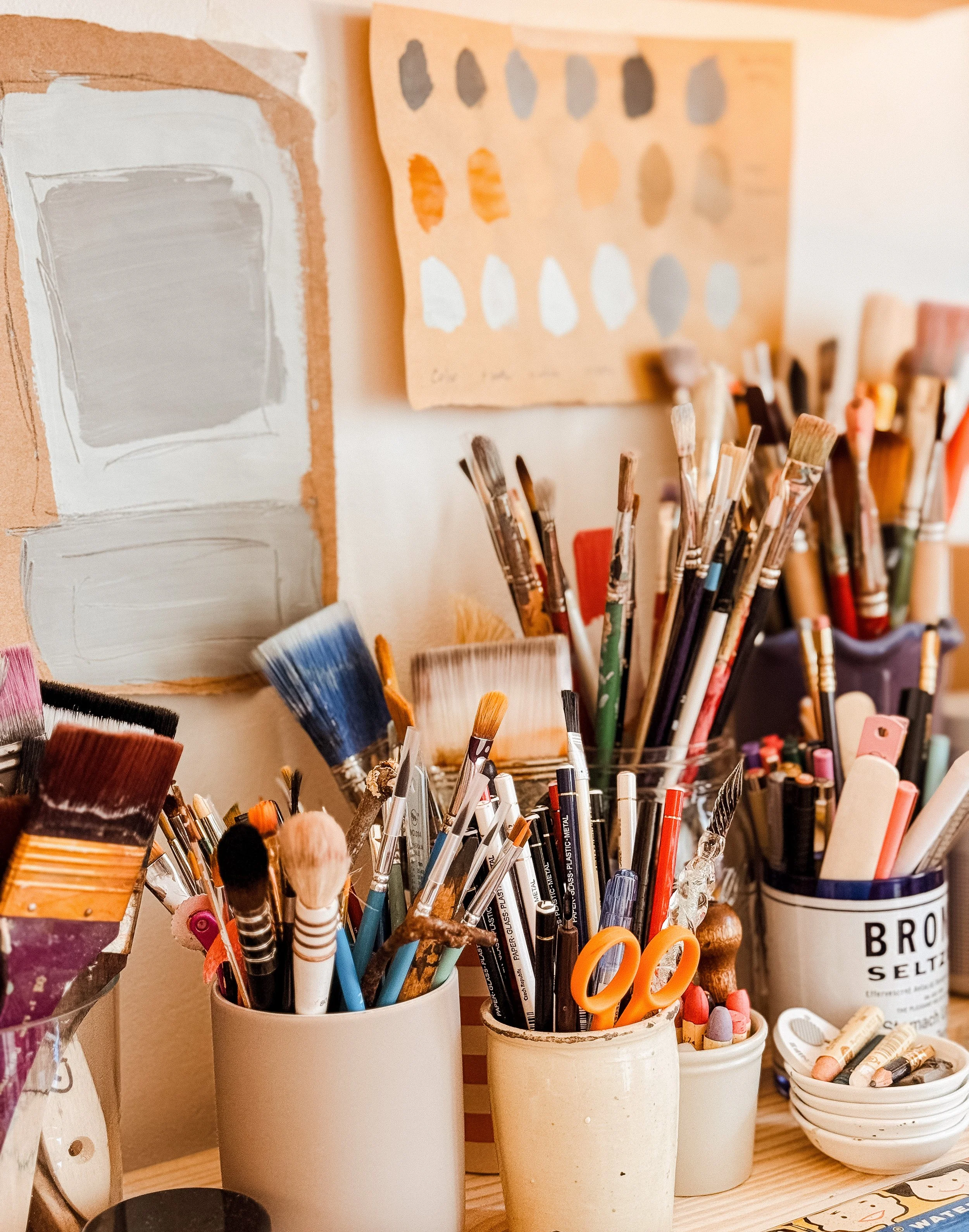 An art workspace with various paintbrushes, pens, and art supplies in cups and containers on a wooden surface, with paintings and color swatches on the wall behind.