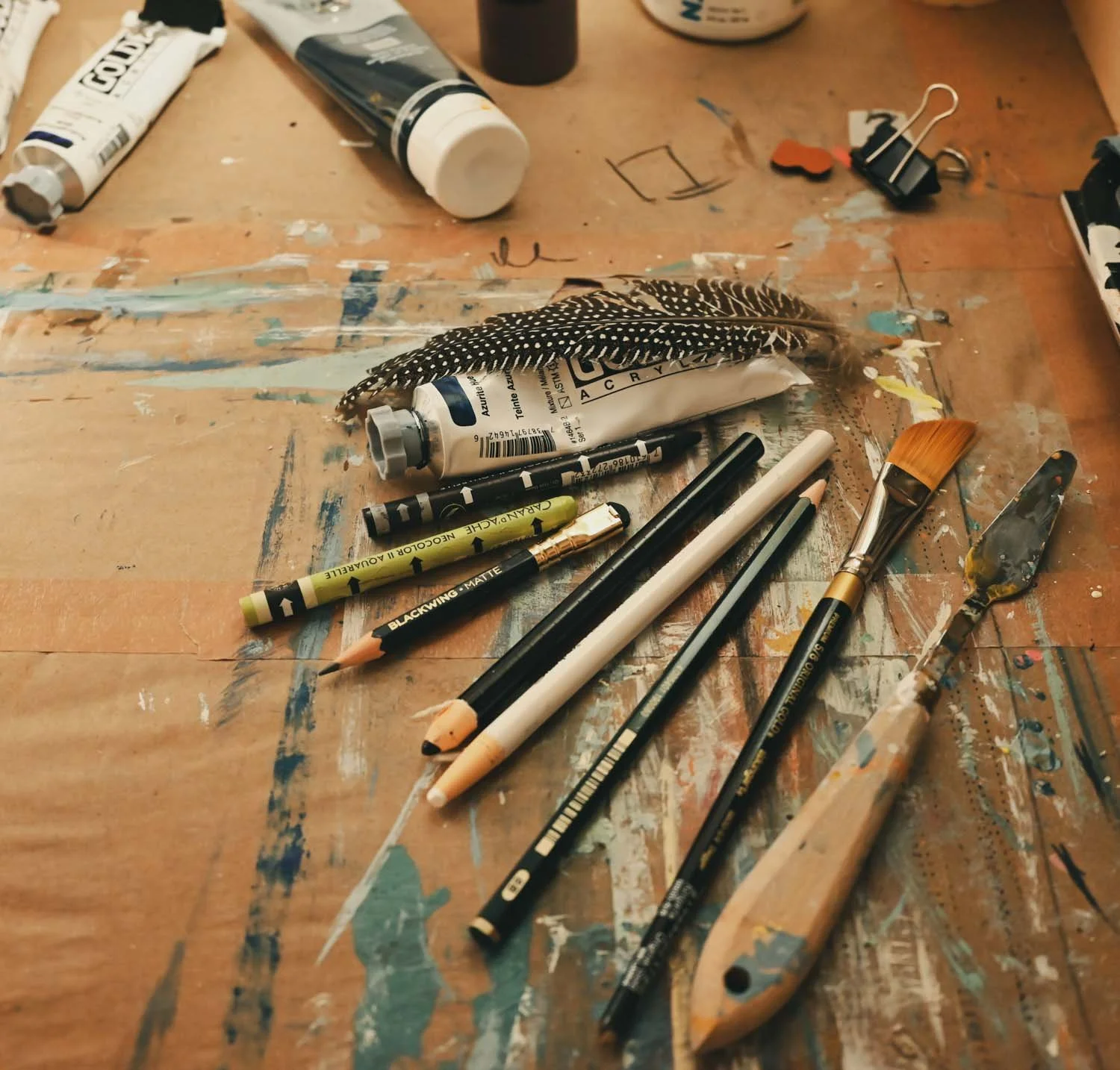 Art supplies on a wooden table, including tubes of paint, paintbrushes, pencils, and miscellaneous tools.