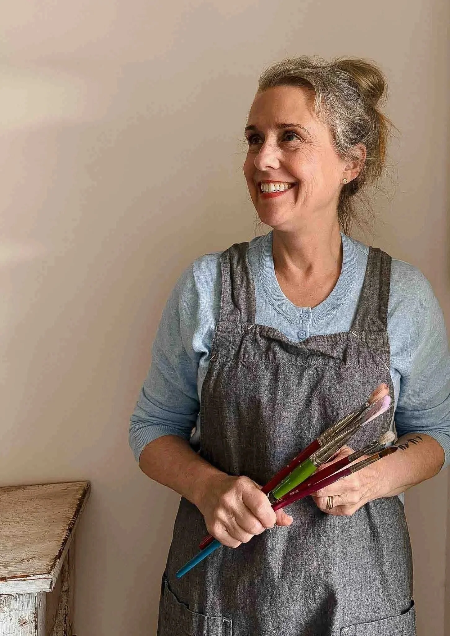 Smiling woman with gray hair in a bun, wearing a light blue shirt and gray apron, holding paintbrushes.