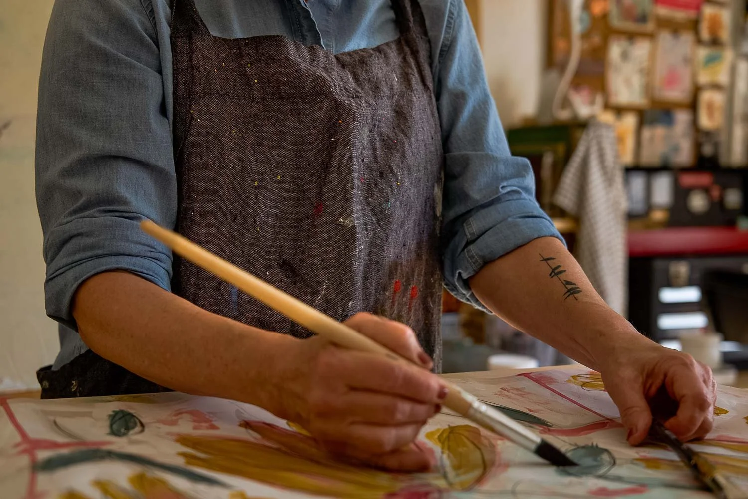 Laura Cantral in a blue shirt and black apron painting on a large piece of paper with paints. She is holding a paintbrush and working on colorful designs. Laura's left forearm has a tattoo of birds on a wire.