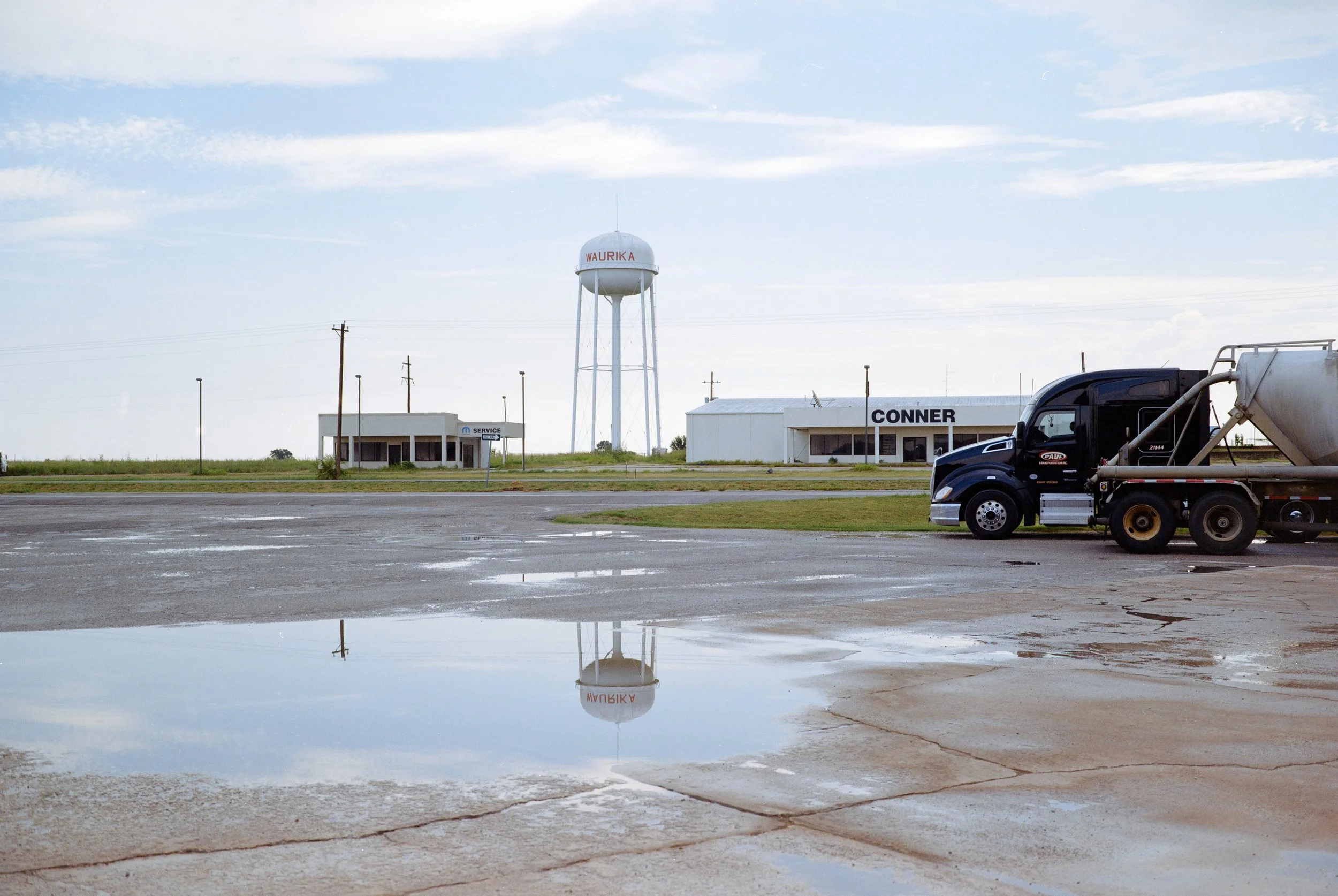 Un parcheggio vuoto con pozzanghere riflettenti, un camion nero sul lato destro, edifici con segni 'SERVICE' e 'CONNER' e a cavallo di un campo aperto con un serbatoio dell'acqua sopra di un palo a torre in lontananza, sotto un cielo parzialmente nuv