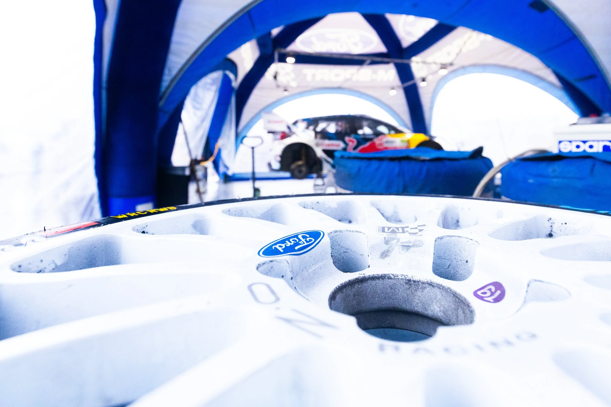 Nei box dei rally, un cerchione bianco in primo piano e una vettura da rally sullo sfondo con strutture in tessuto blu.