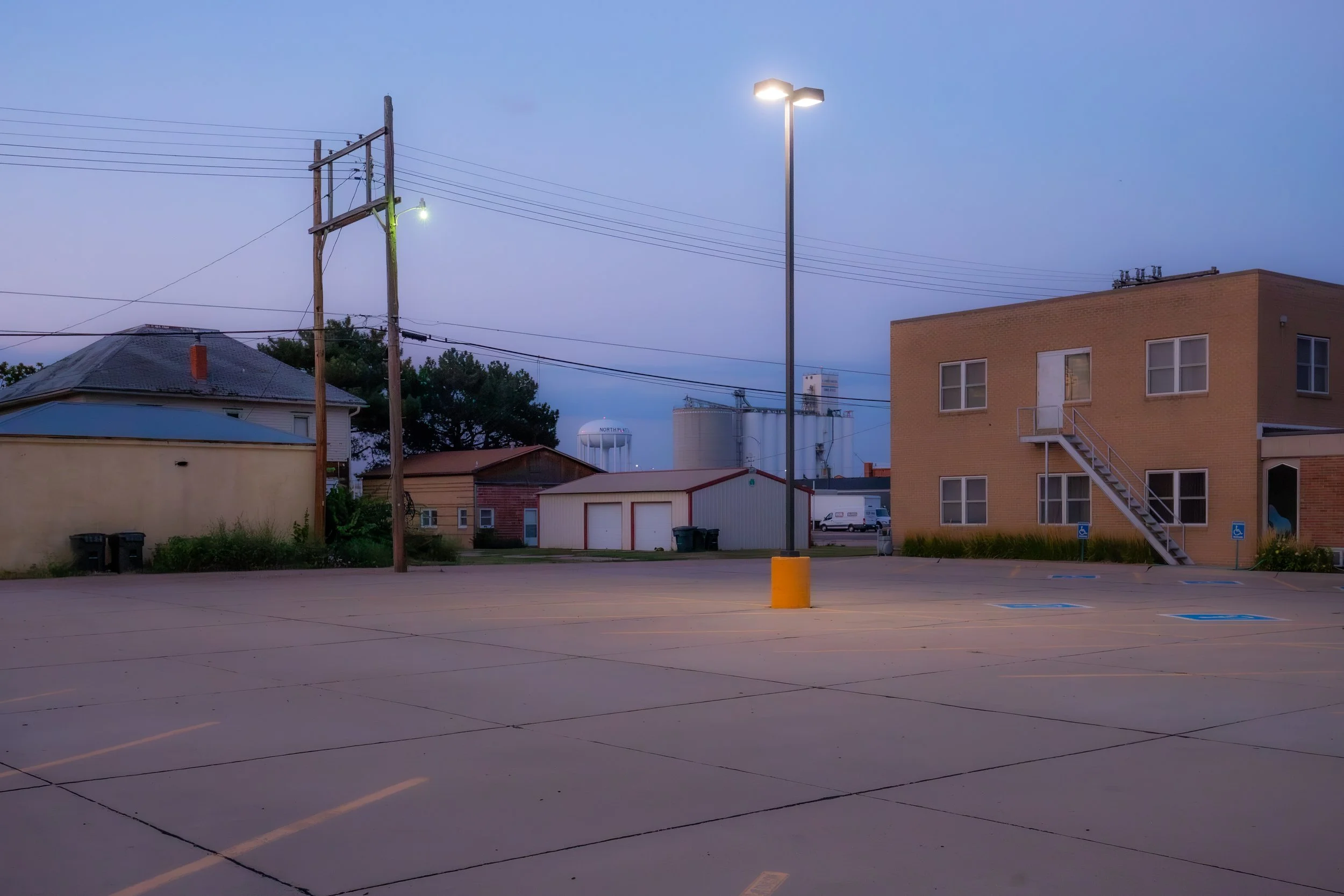 Un parcheggio vuoto con alcune aree riservate ai disabili, un edificio giallo con scala esterna e illuminazione pubblica, al tramonto o all'alba.